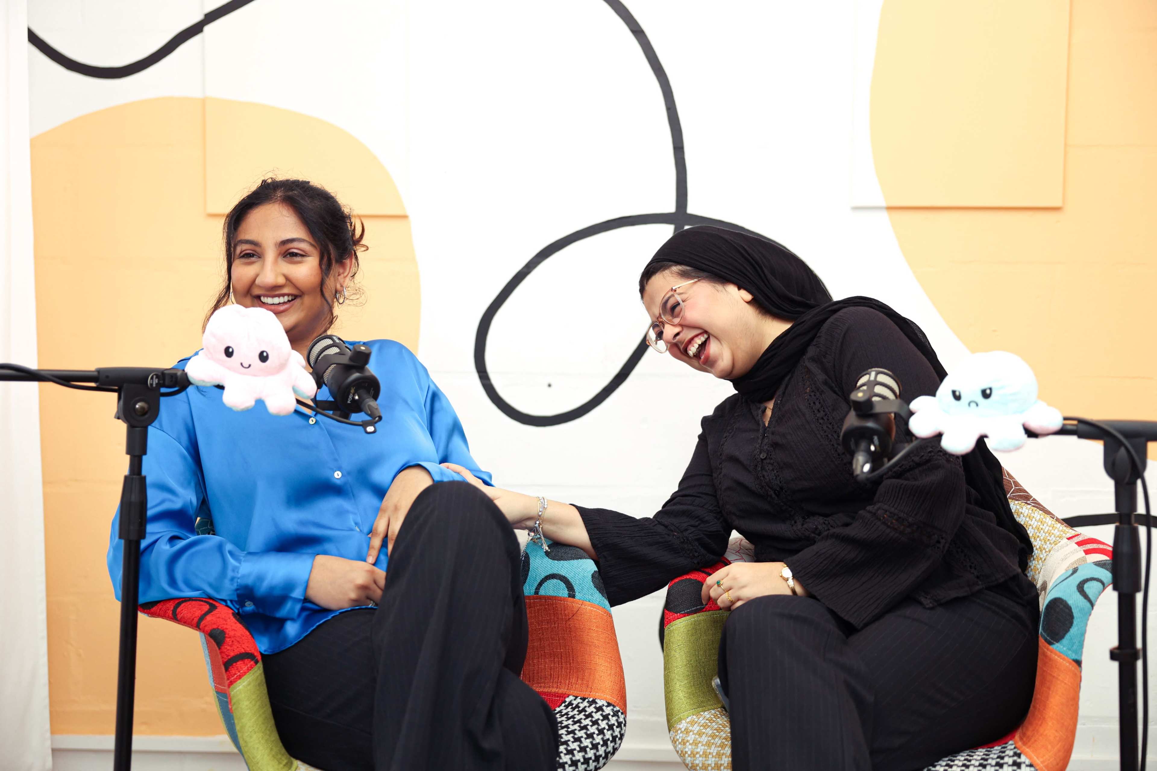 Two women sit in colorful chairs, laughing together in a podcast recording studio with microphones and plush octopus decorations nearby.
