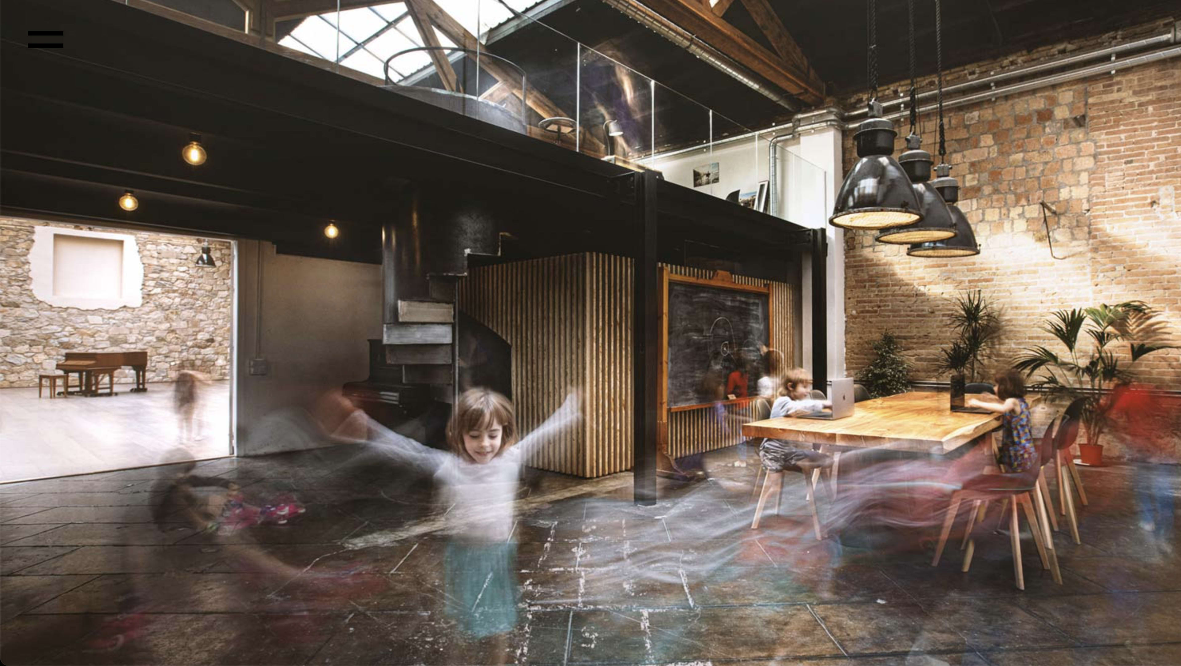 A spacious open-concept room features a dining table surrounded by chairs, a wooden staircase, and children playing in motion, with large windows allowing natural light to illuminate the space.
