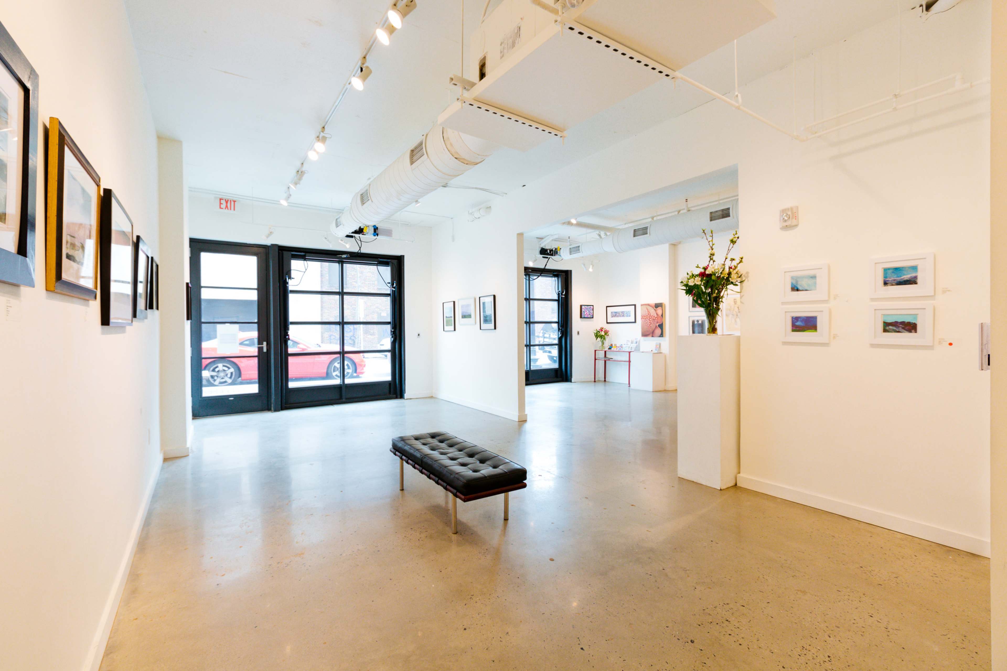 An art gallery featuring white walls, a polished concrete floor, and a central black bench with framed artwork displayed around the space.