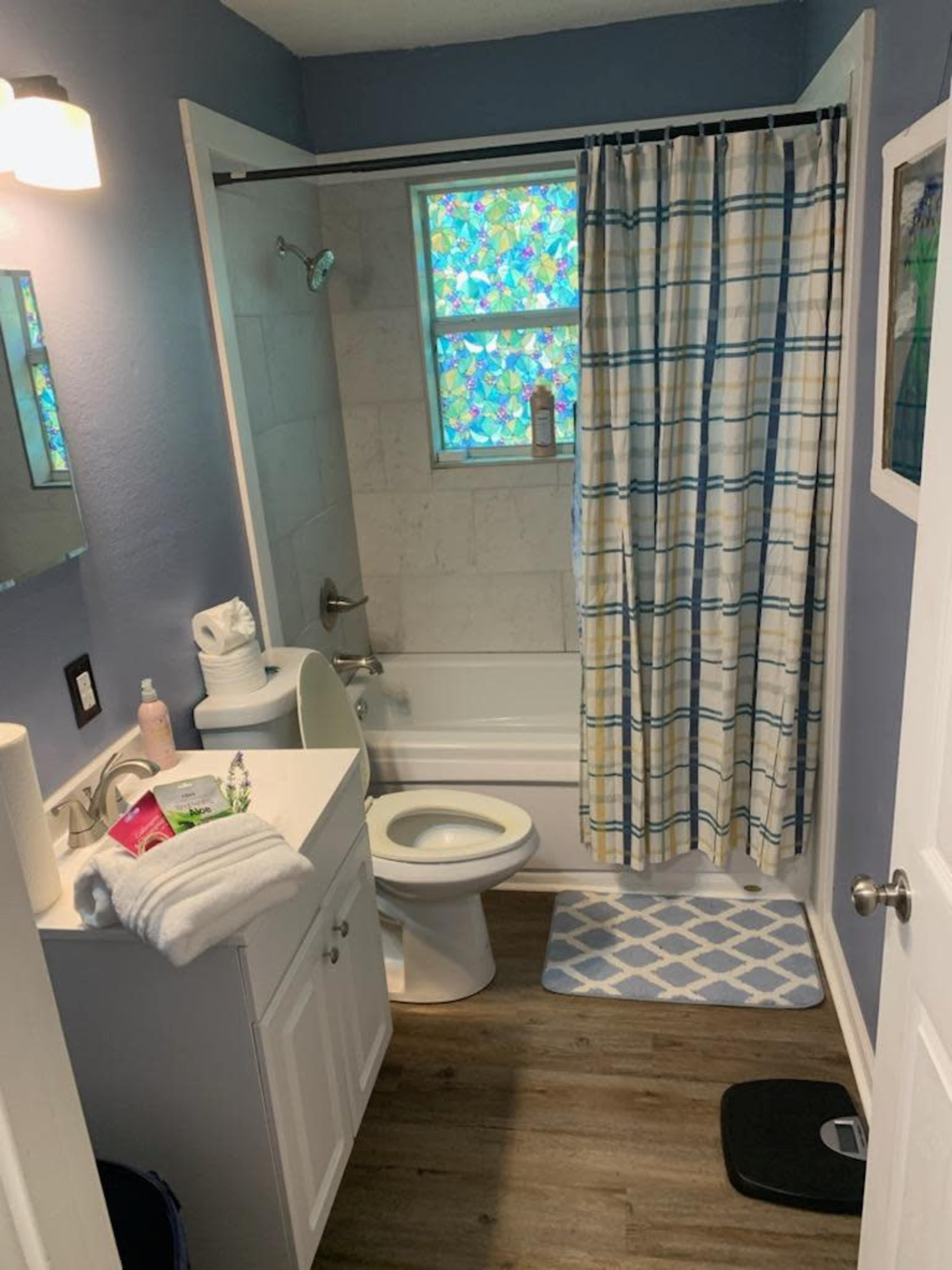 The image shows a bathroom with a shower, a toilet, a sink, and a window, featuring a colorful shower curtain and a bath mat on the floor.