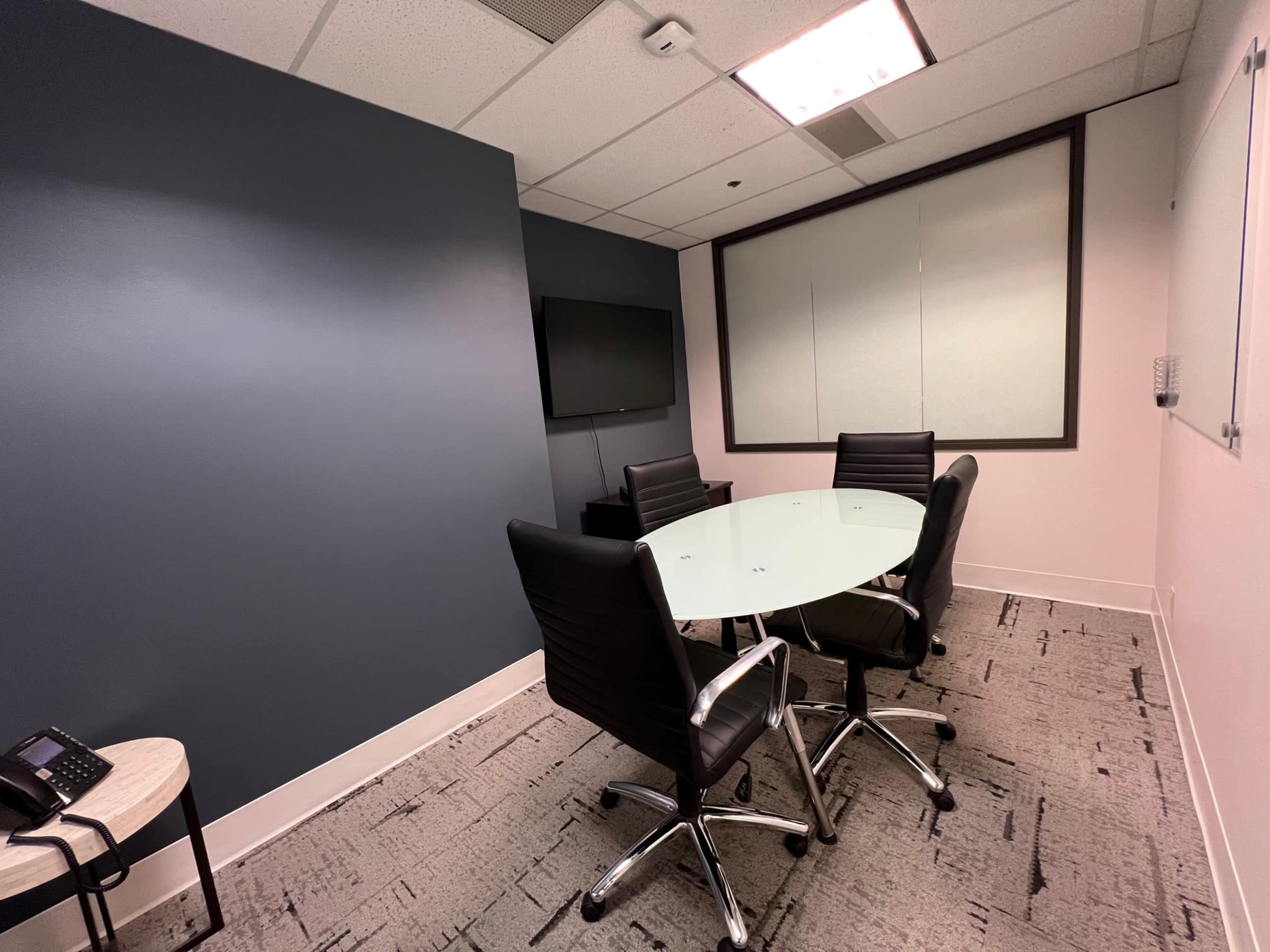 A small conference room features a round glass table surrounded by four black chairs, with a wall-mounted television and a whiteboard.