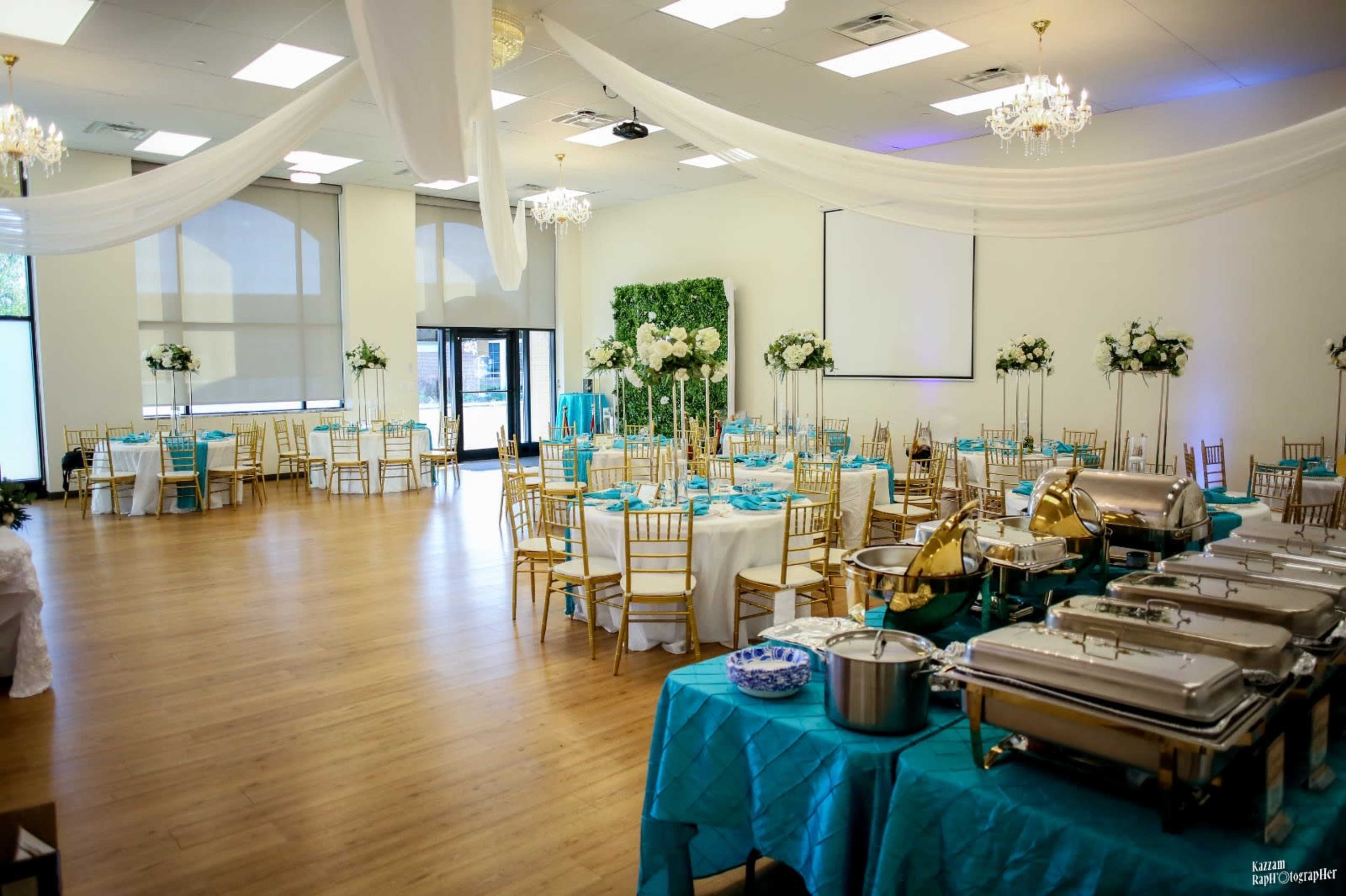 The image shows a banquet hall set up for an event, featuring round tables with white tablecloths and turquoise accents, surrounded by gold chairs, and a buffet area with metal food warmers in the foreground.