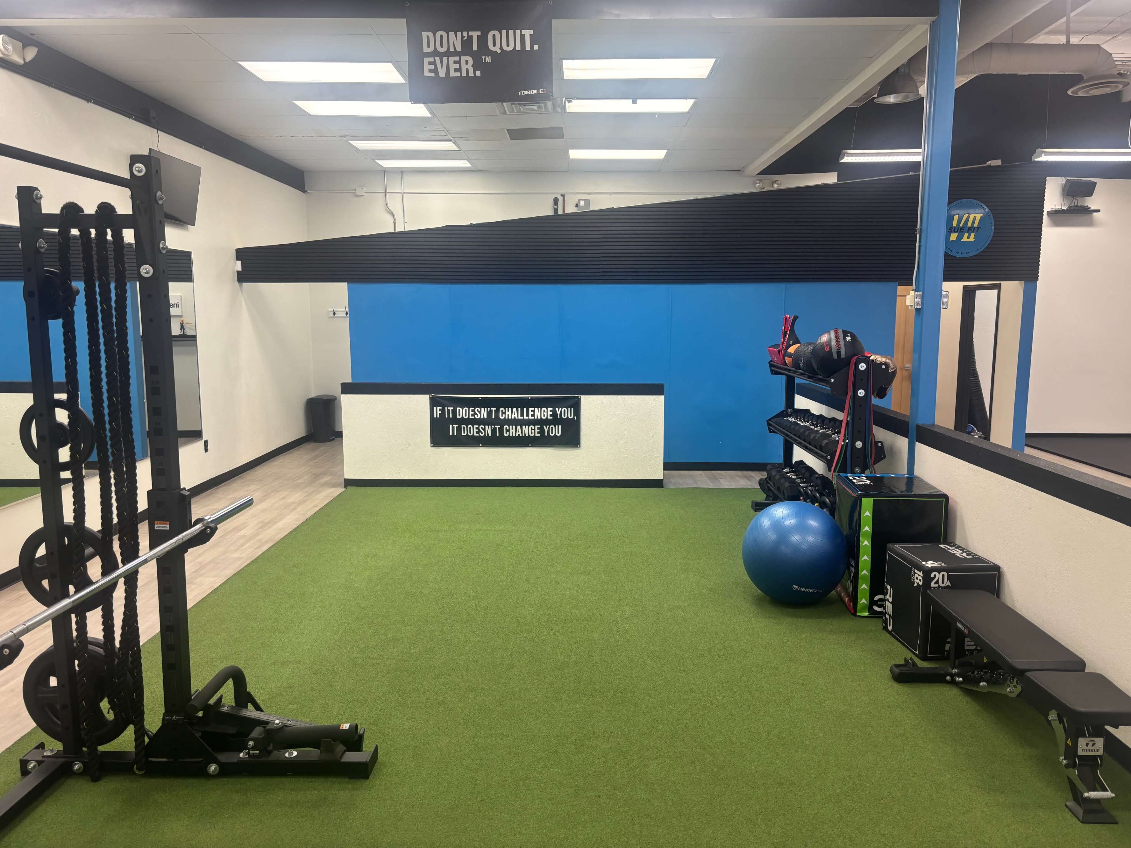 The image shows a modern gym interior with green turf flooring, various exercise equipment, and motivational signage on the wall.