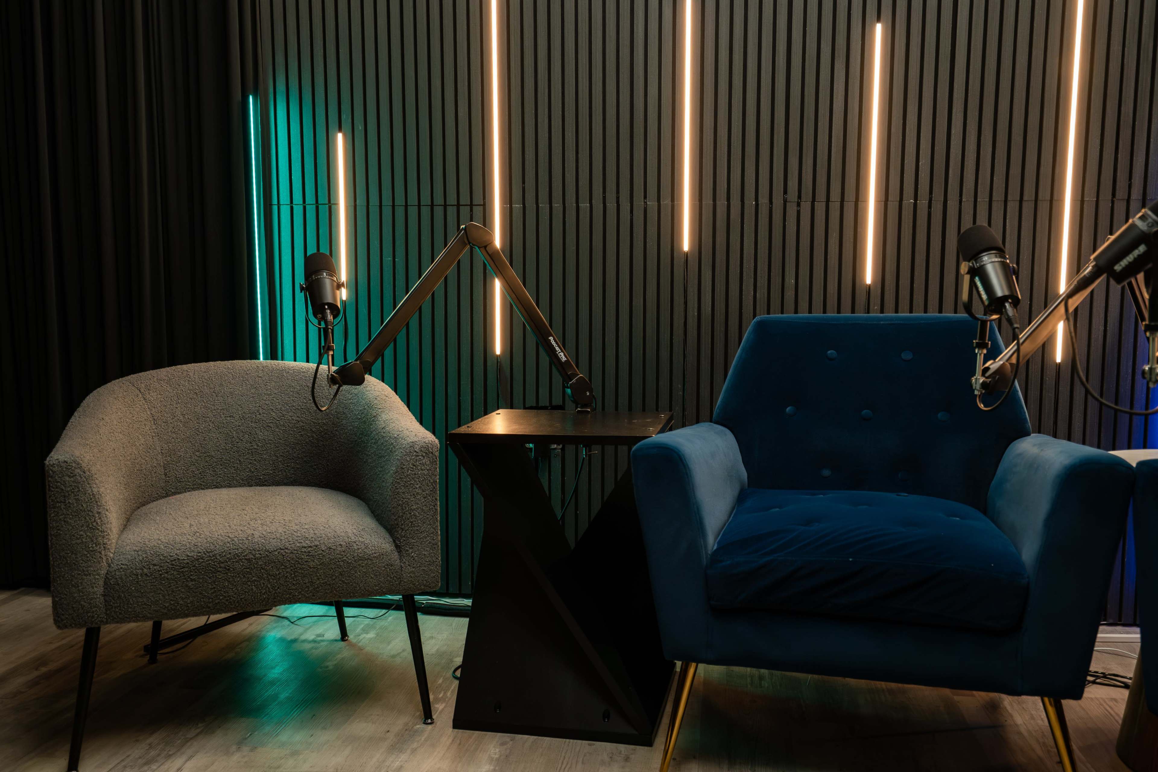 The image shows a podcast studio setup with two chairs, a small black table, and microphones mounted on adjustable arms against a backdrop of vertical wooden panels and illuminated strips.