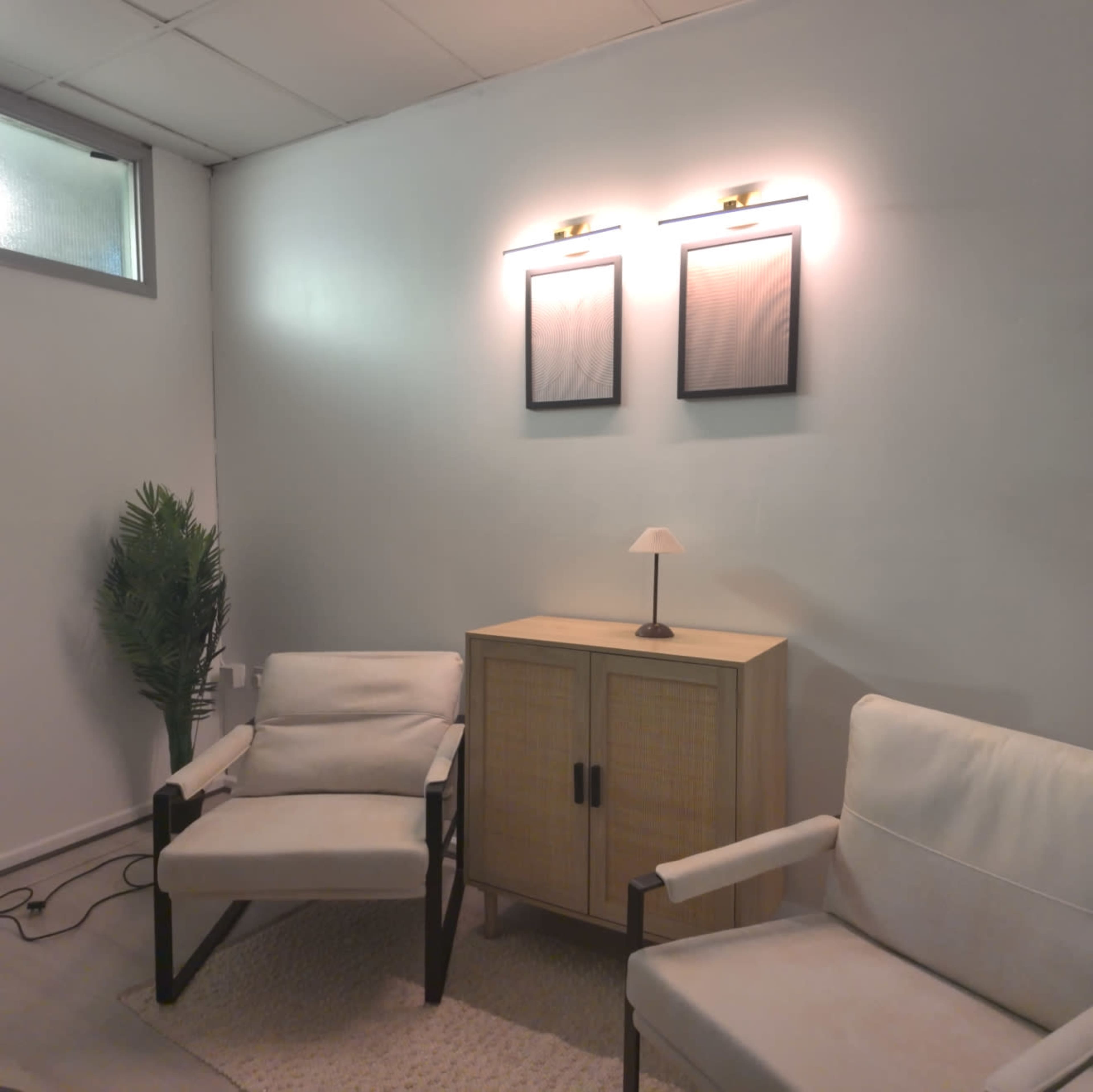 The image shows a minimalist living space featuring two armchairs, a small lamp on a cabinet, and framed artworks on the wall, with a potted plant in the corner.