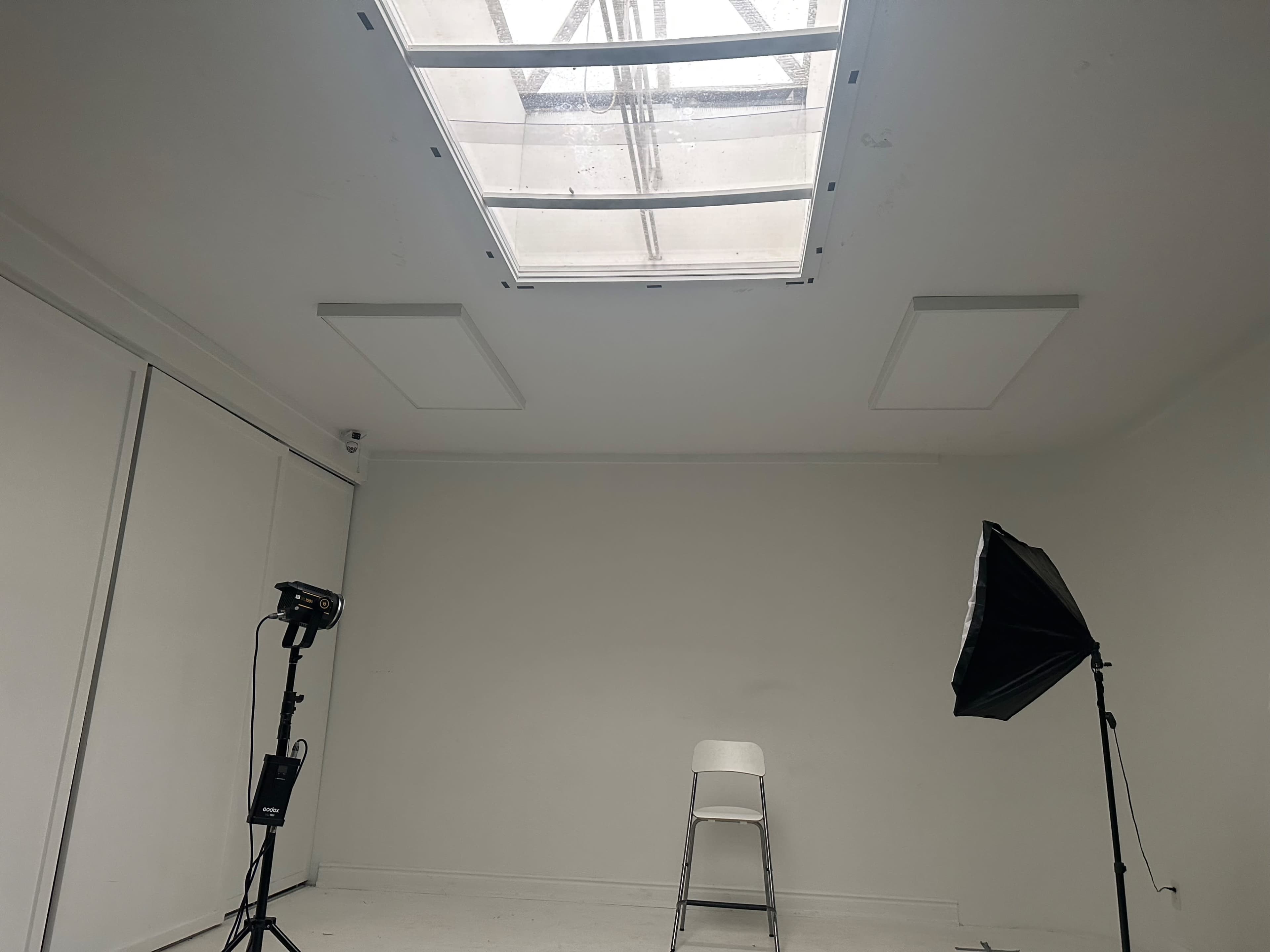 The image shows a minimalist white room with a skylight, a folding chair in the center, and a lighting setup consisting of a stand and a softbox.