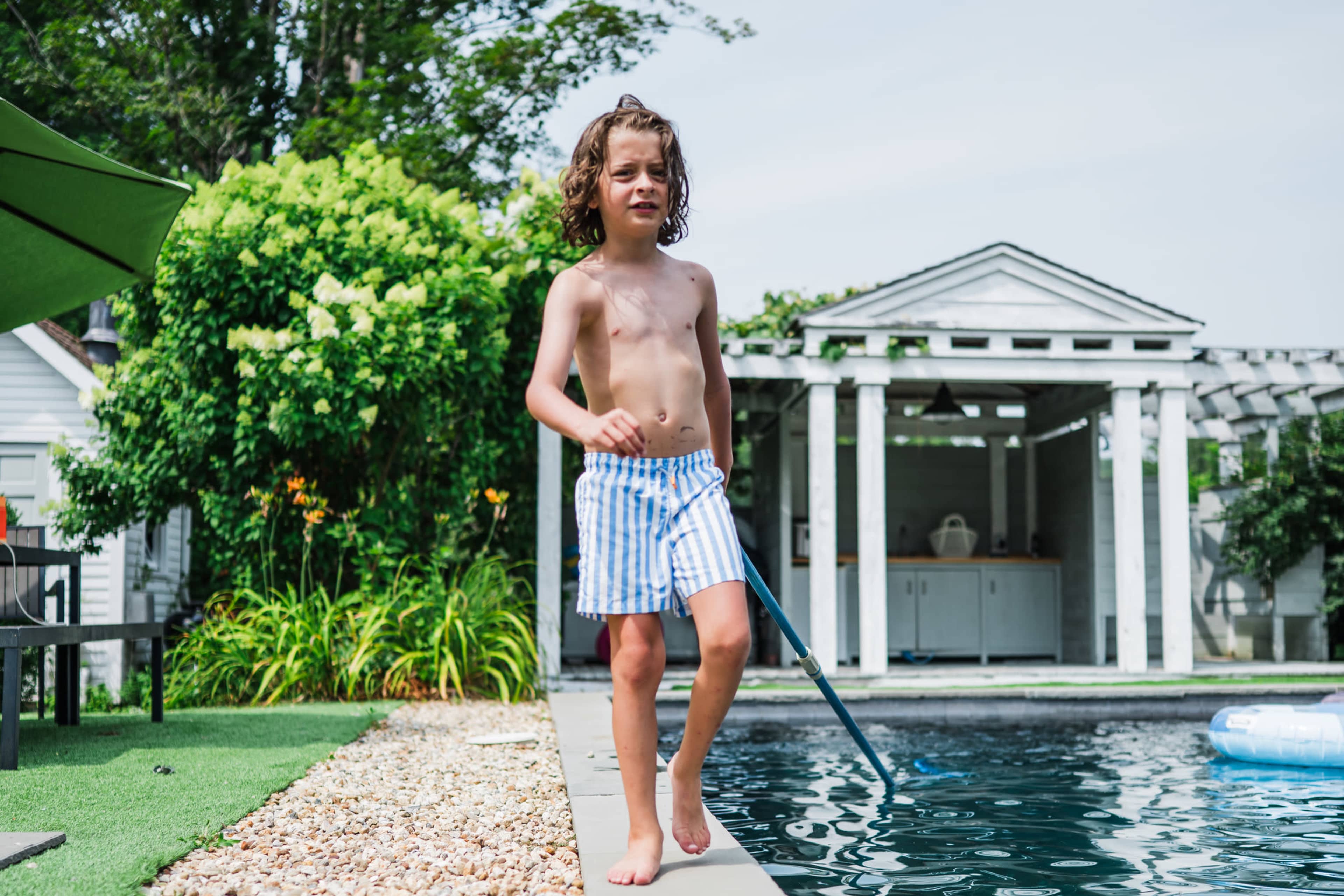 A boy with curly hair in striped swim shorts walks along the edge of a swimming pool.
