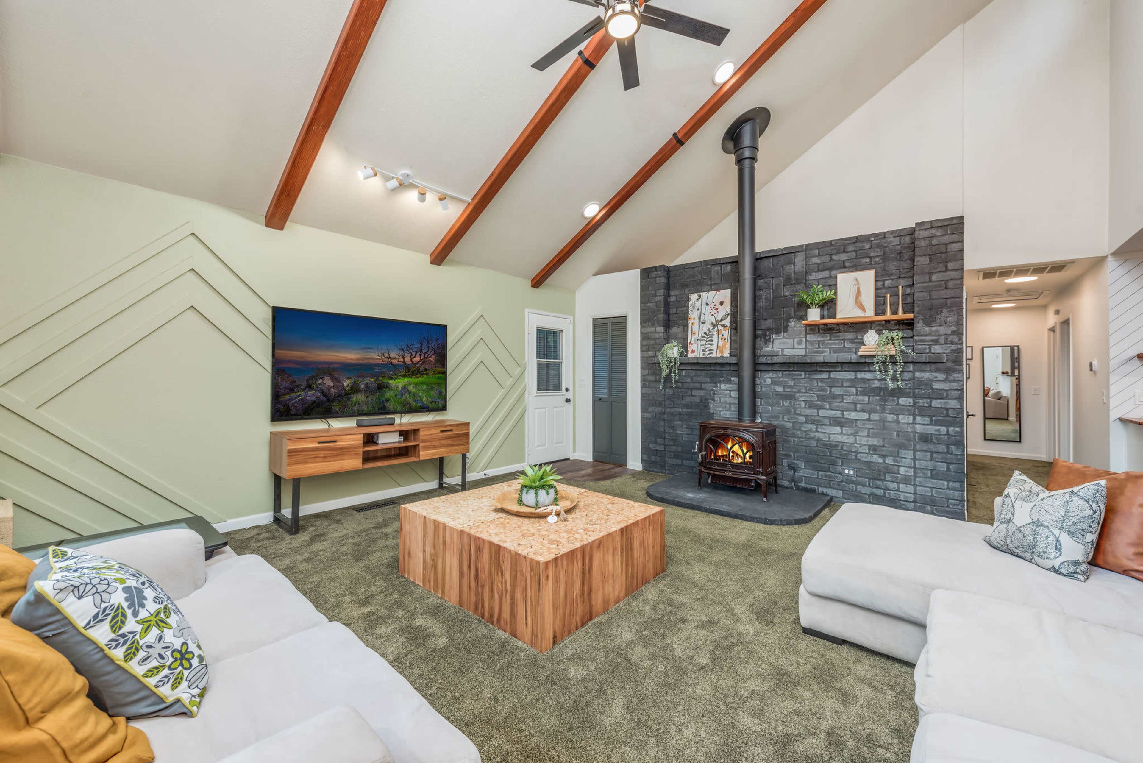 The living room features a wood-burning stove, a large coffee table, and a TV mounted on the wall, with vaulted ceilings and a mix of green and gray decor.