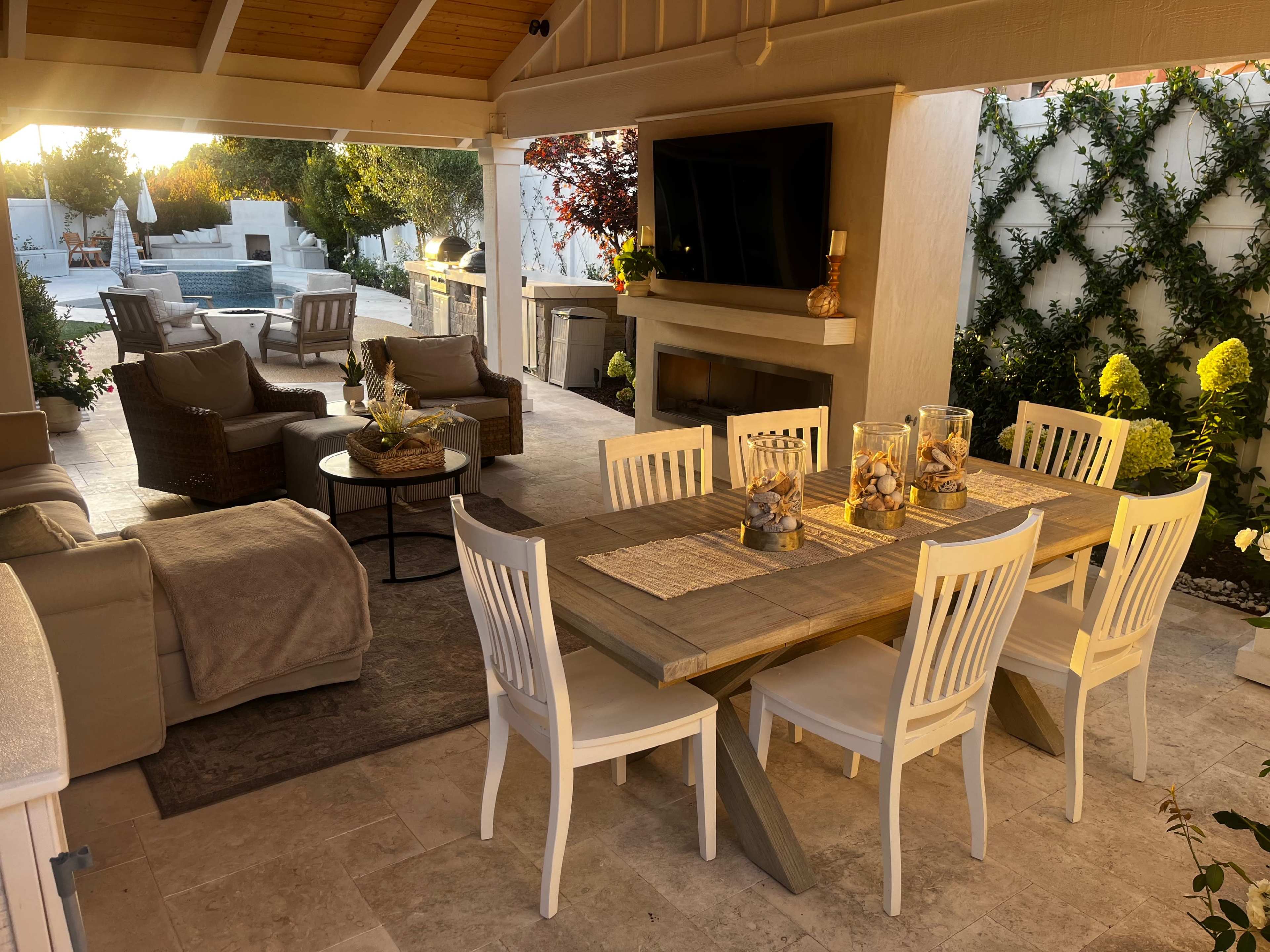 A modern outdoor patio with a dining table, comfortable seating, and a television, illuminated by warm sunset light.