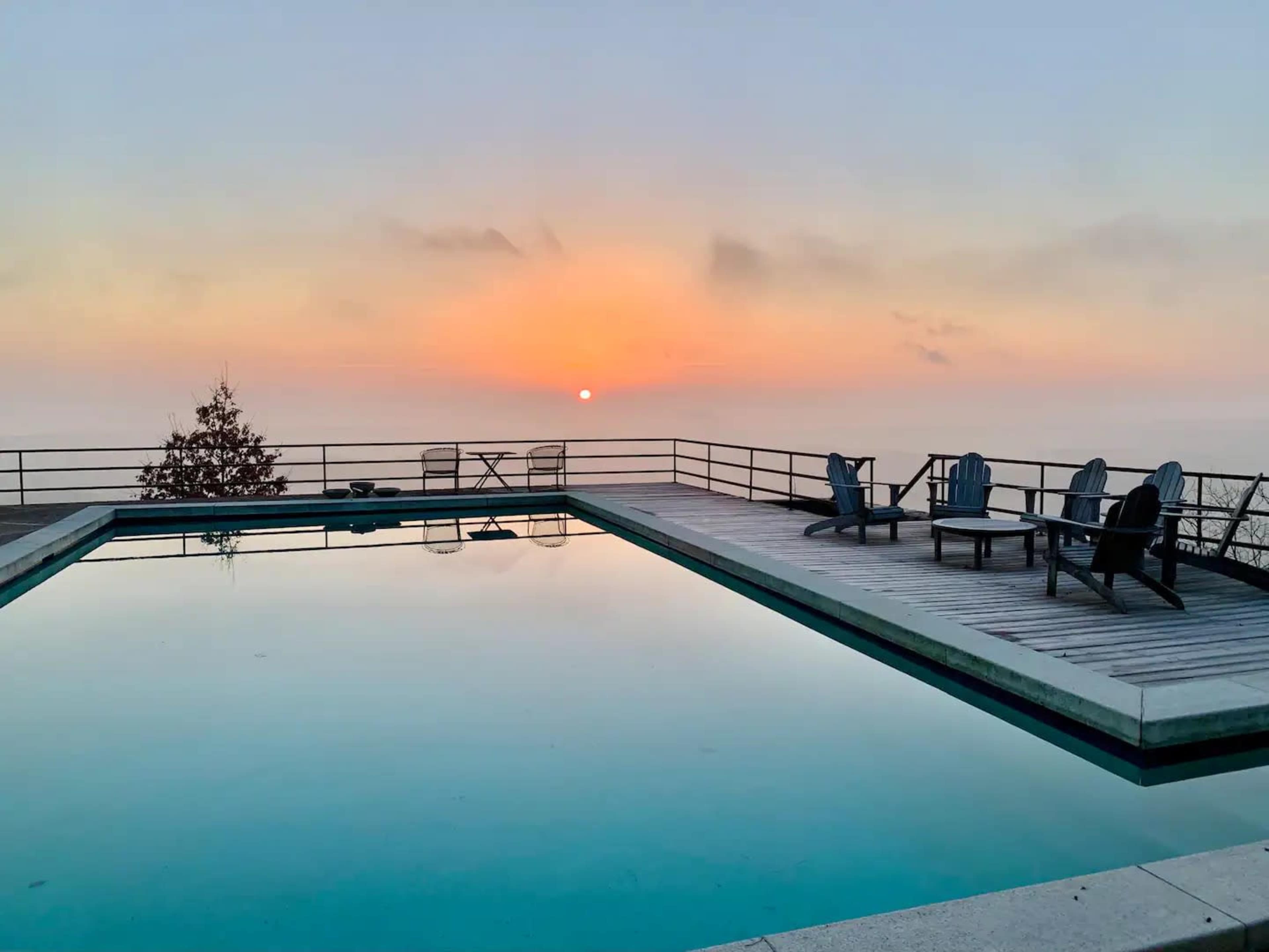 The scene features a rectangular swimming pool reflecting a sunset, with several wooden lounge chairs positioned nearby against a backdrop of a hazy horizon.