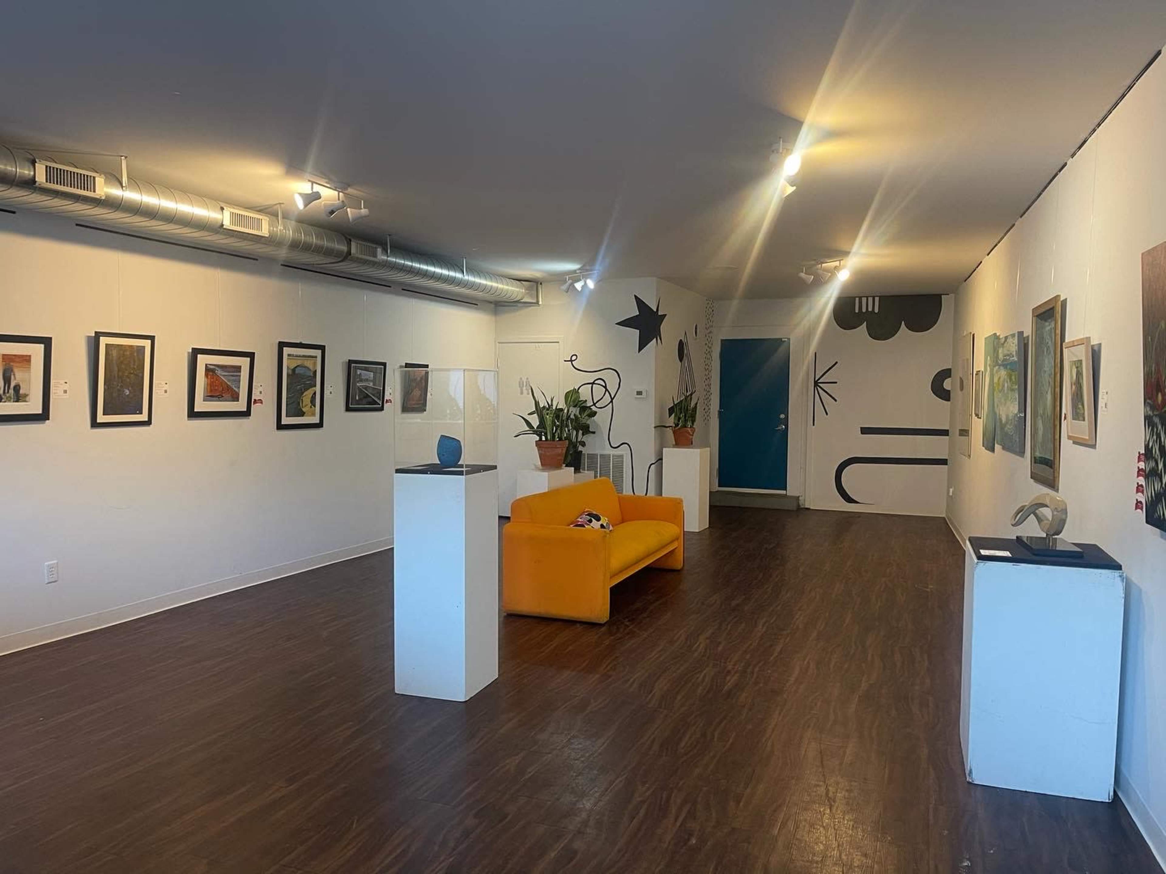 An art gallery with framed artworks on the walls, a bright orange couch in the center, and a few indoor plants near the seating area.