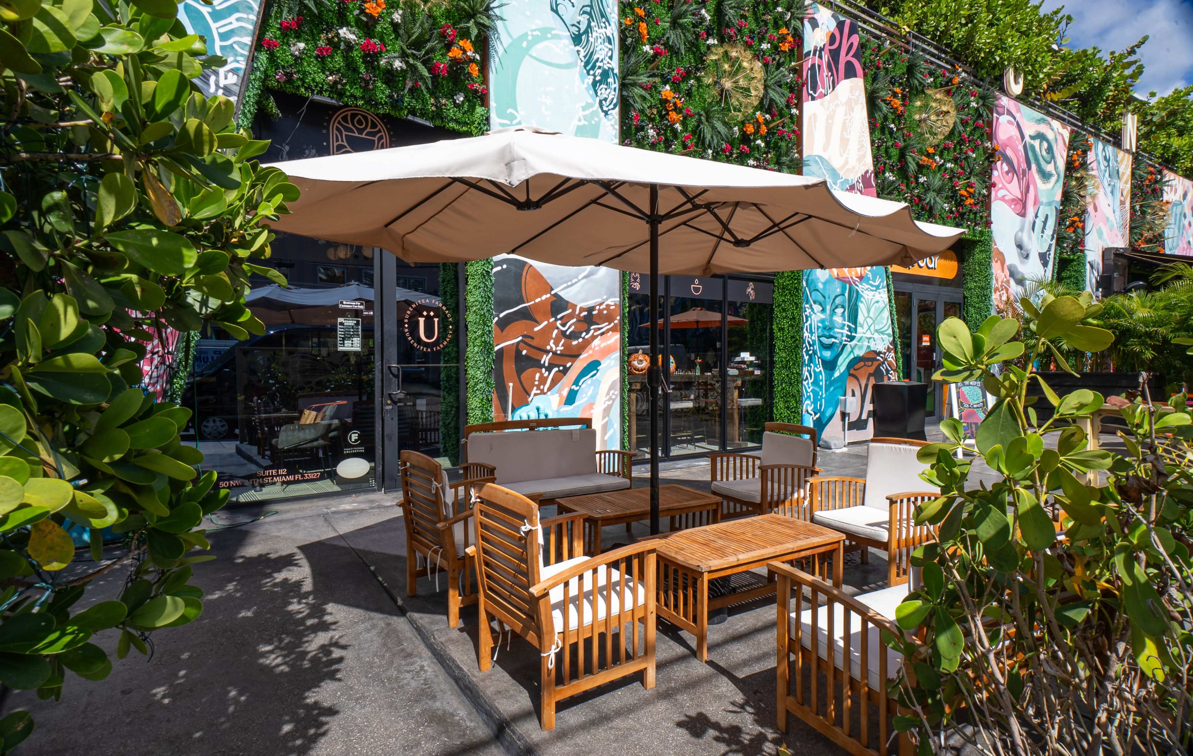 The image shows a partially shaded outdoor seating area at a restaurant, featuring wooden furniture and vibrant murals on the walls.