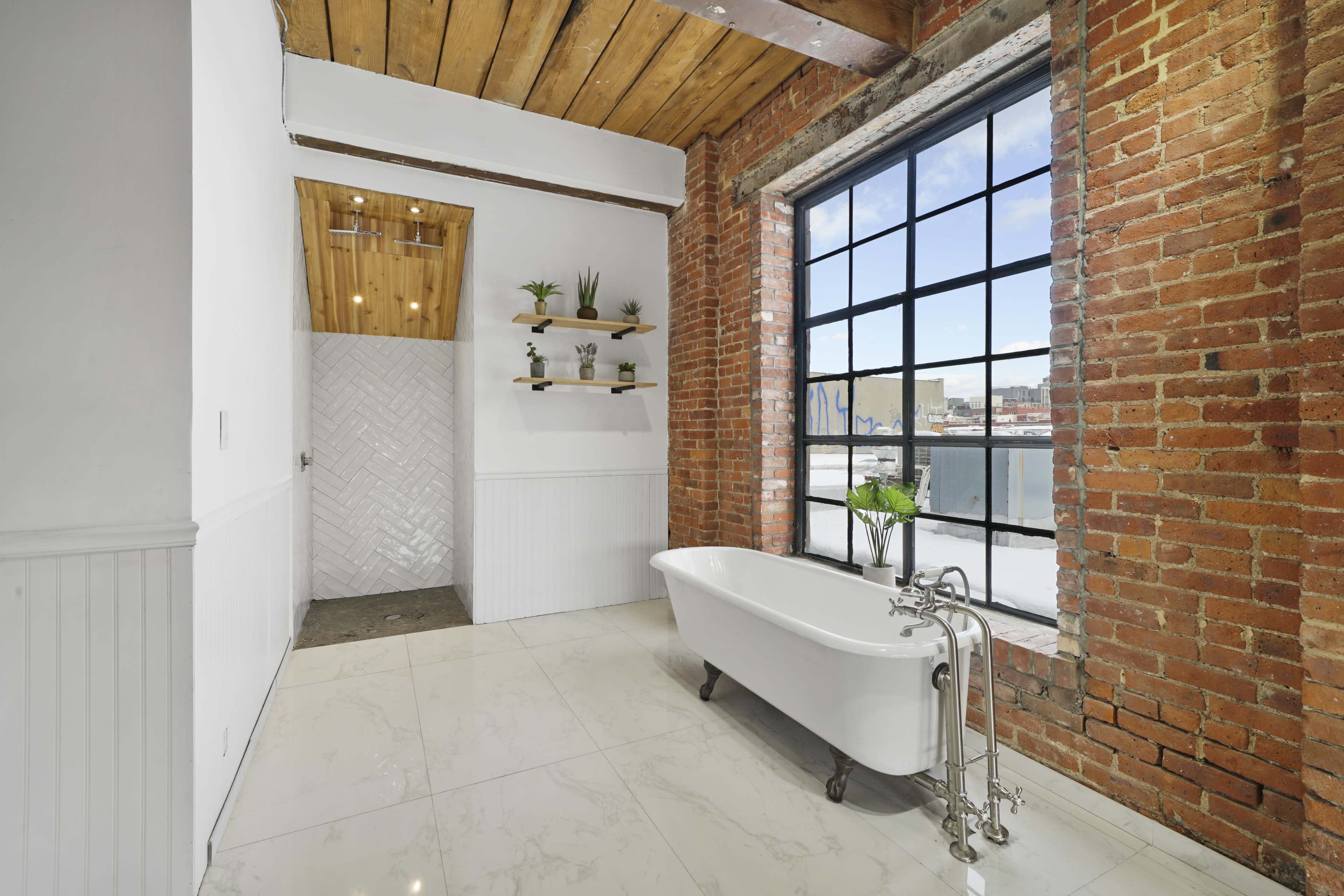 A modern bathroom with a freestanding tub, exposed brick walls, large windows, and a wood-beamed ceiling.