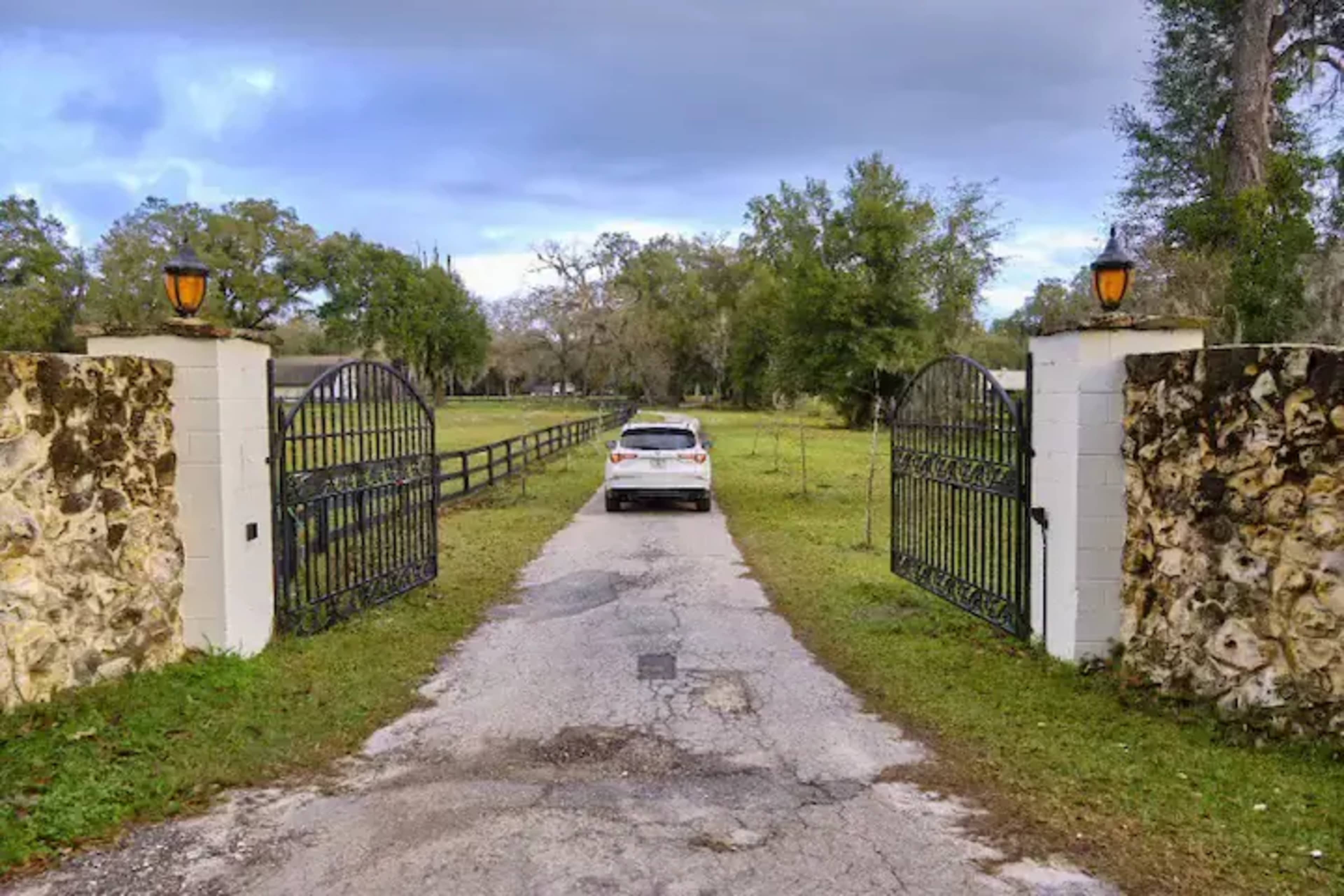 A white car is parked on a gravel driveway leading through wrought-iron gates into a green pasture.