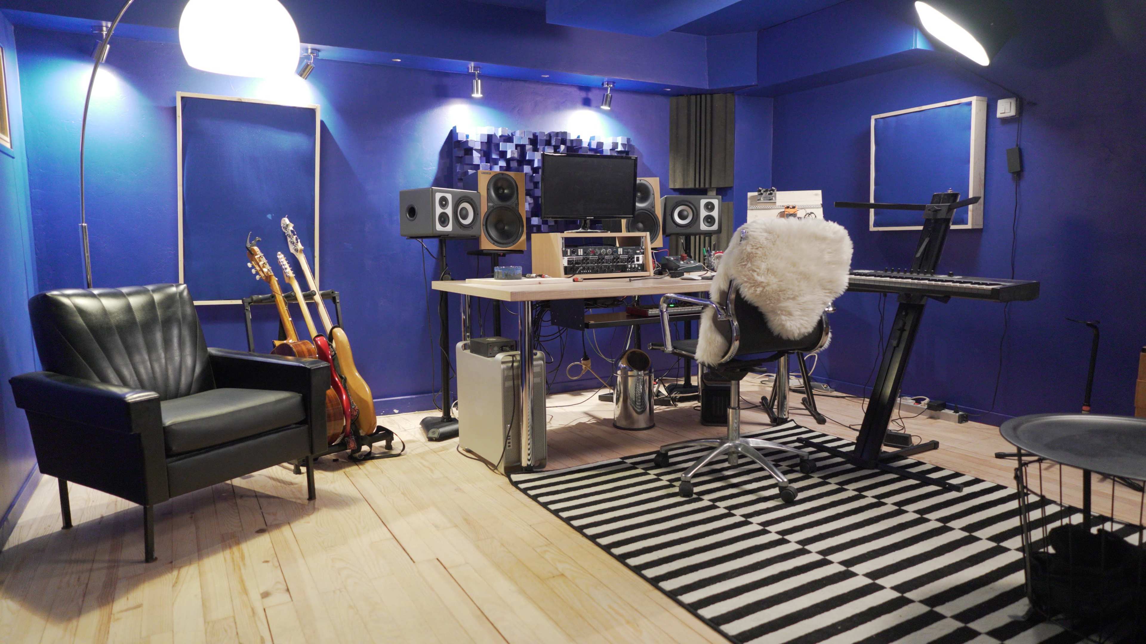 The image shows a music studio with a blue wall, a desk equipped with audio equipment, a keyboard, a black chair, several guitars on stands, and a black and white striped rug.