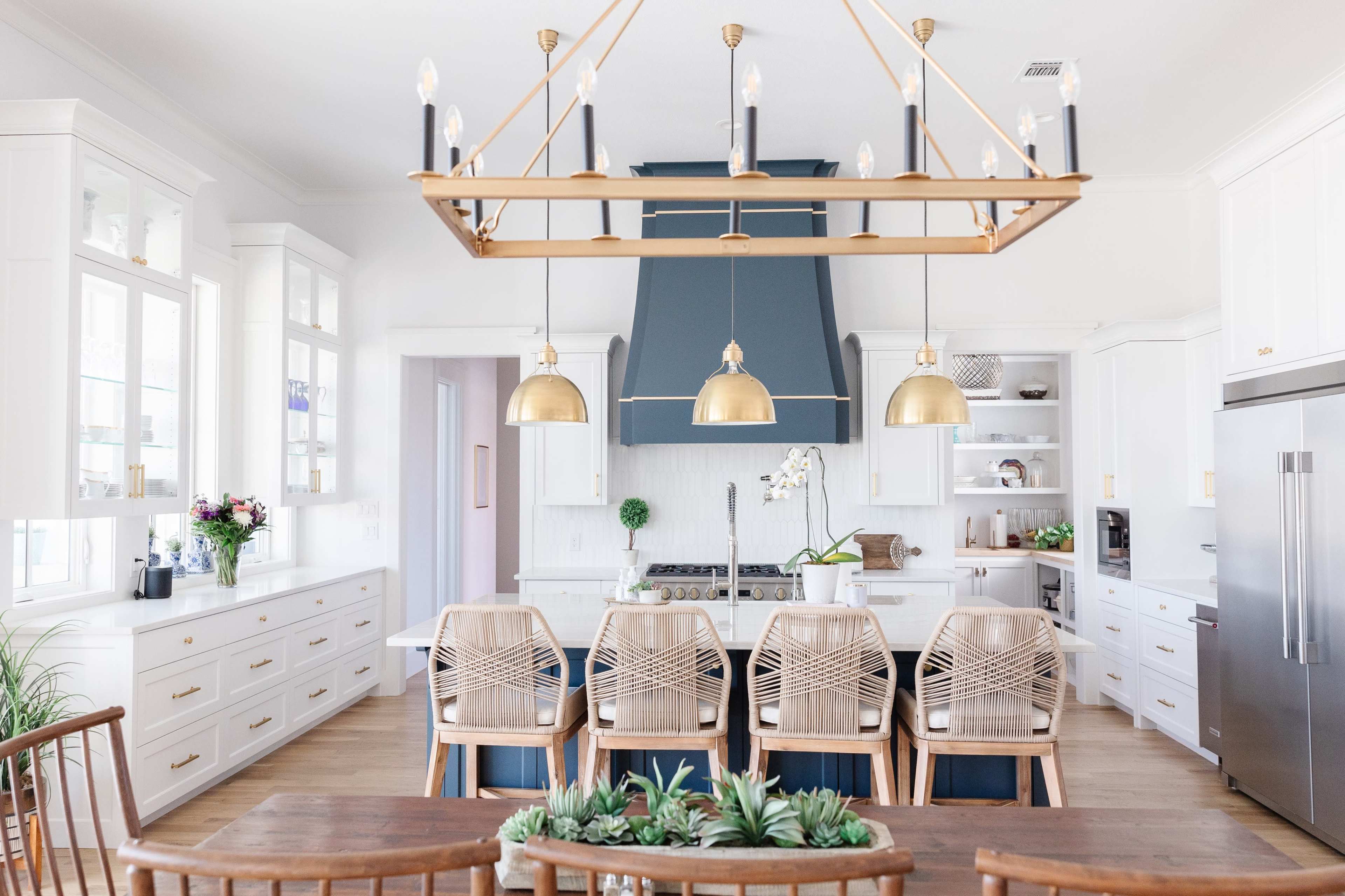 A modern kitchen features white cabinetry, a large central island with pendant lighting, and a dining area with rattan chairs.