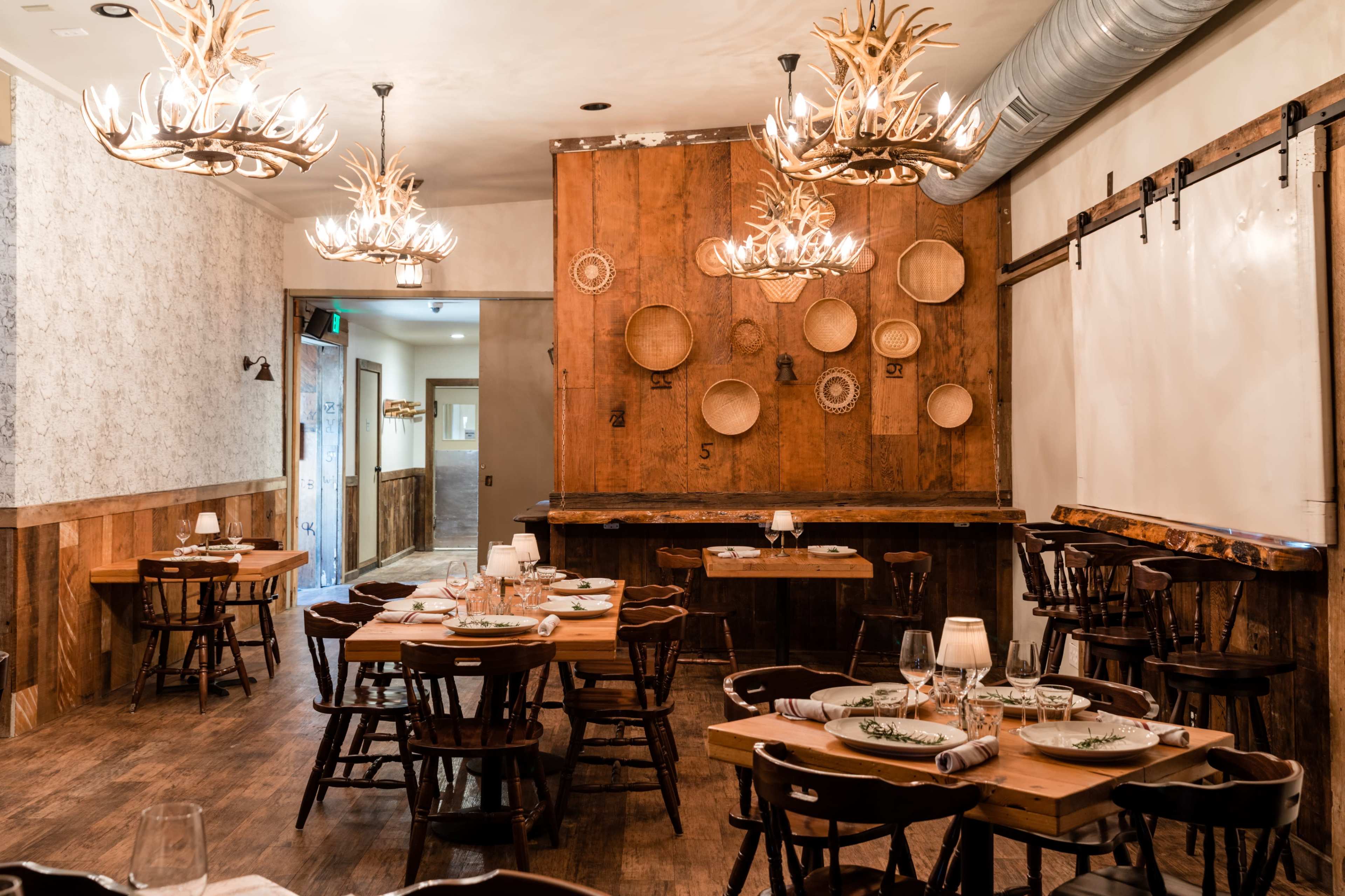 A rustic restaurant interior with wooden tables, chairs, and antler chandeliers, featuring wall decorations of woven baskets.