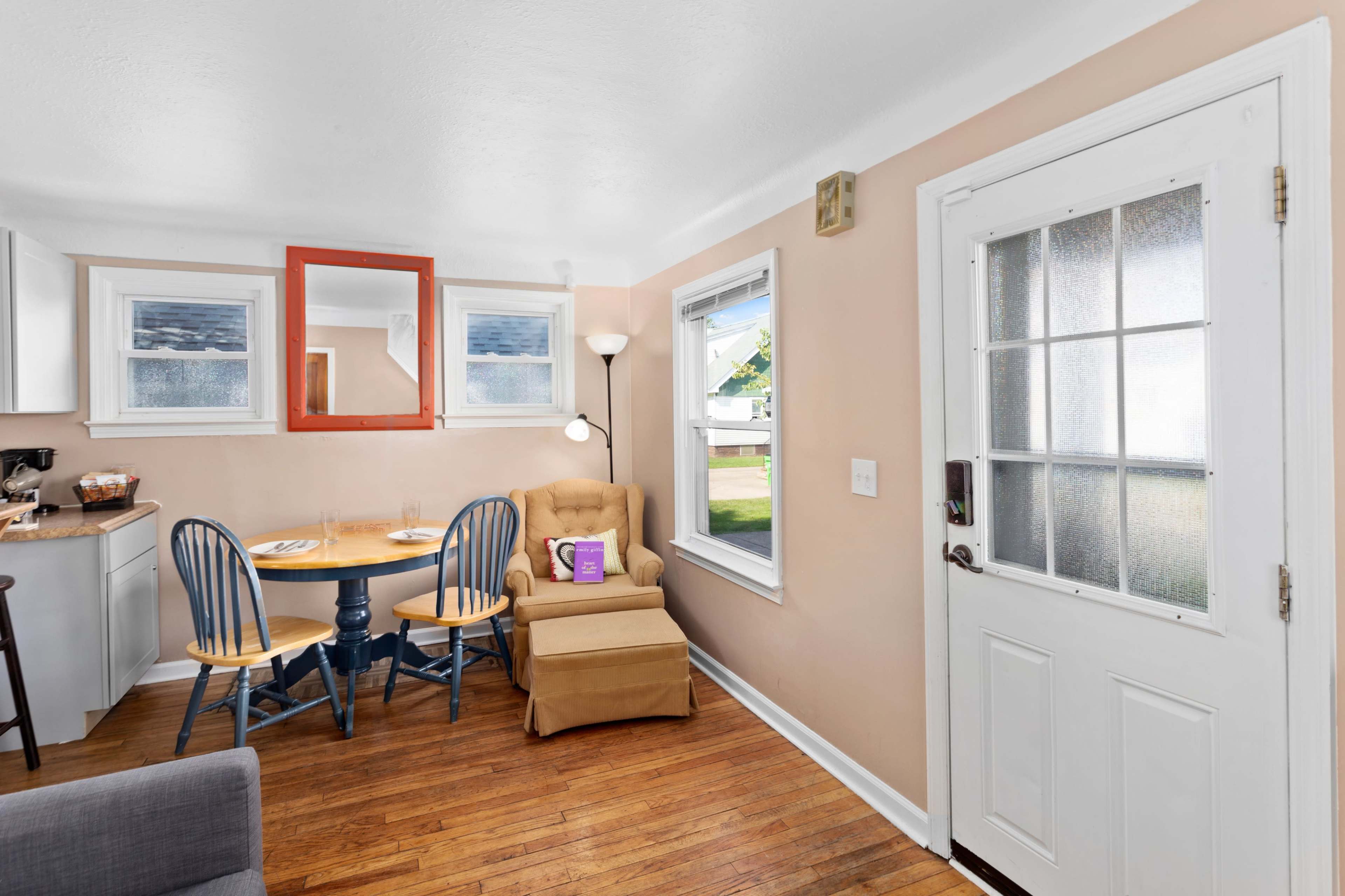 A cozy room features a small dining table with blue chairs next to a yellow armchair, with a door leading outside and a mirror on the wall.