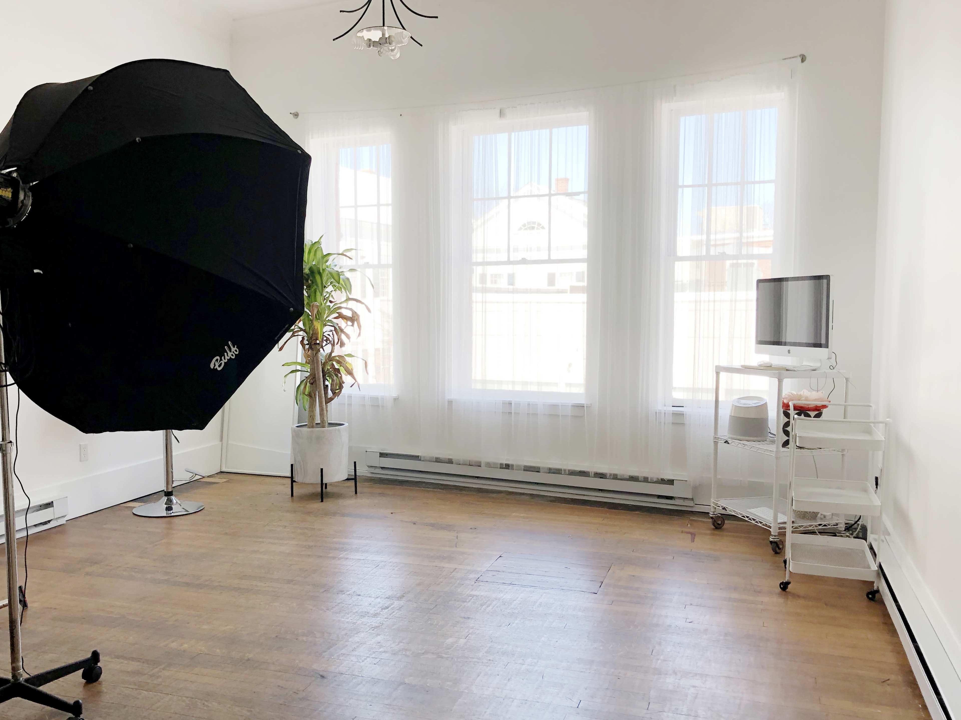 A bright, empty room with large windows, showcasing a photography umbrella, a potted plant, and a small cart with a television.