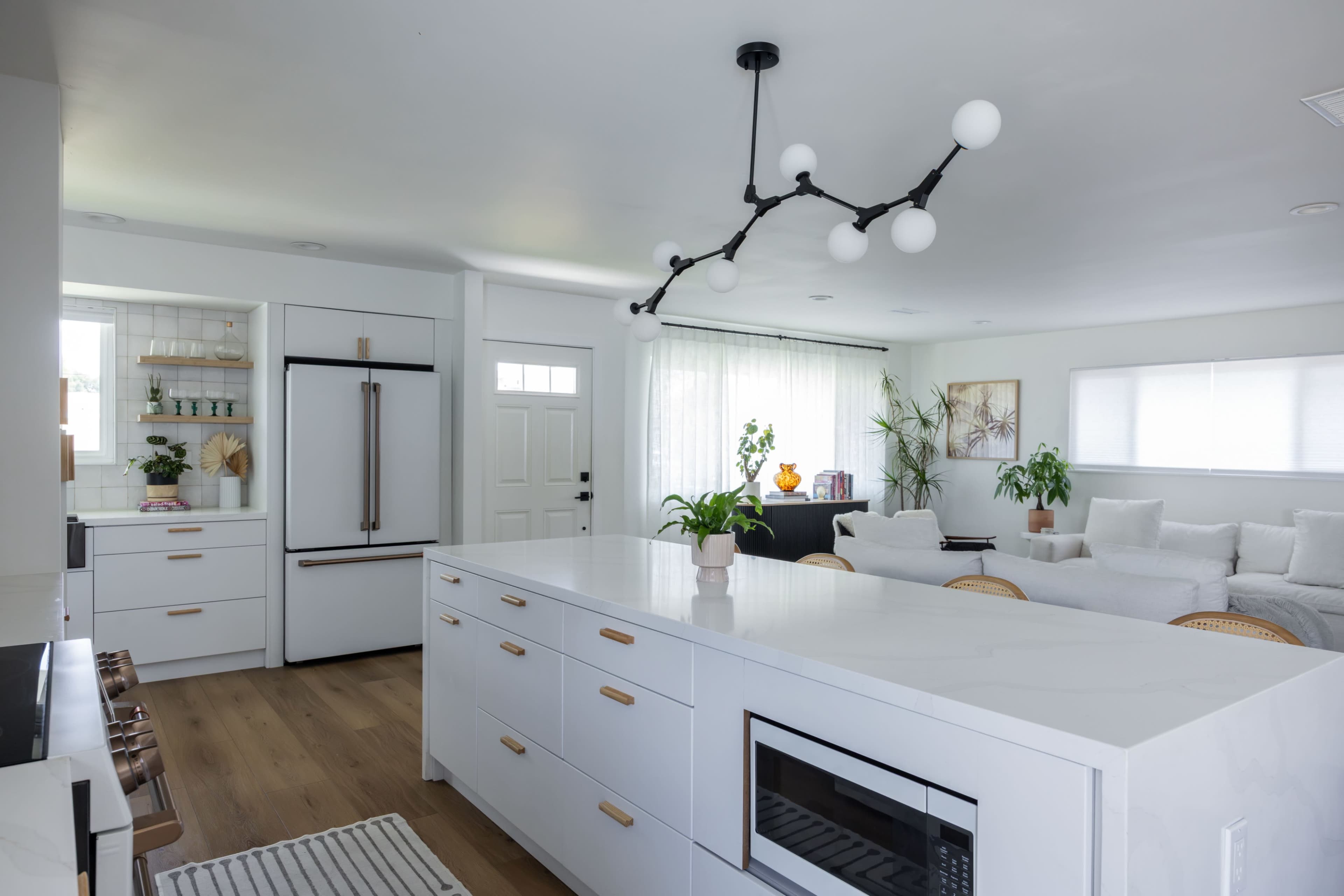 A modern kitchen features a large white island with a sink and microwave, a sleek refrigerator, and an open layout connecting to a sunny living area with plants and a cozy sofa.