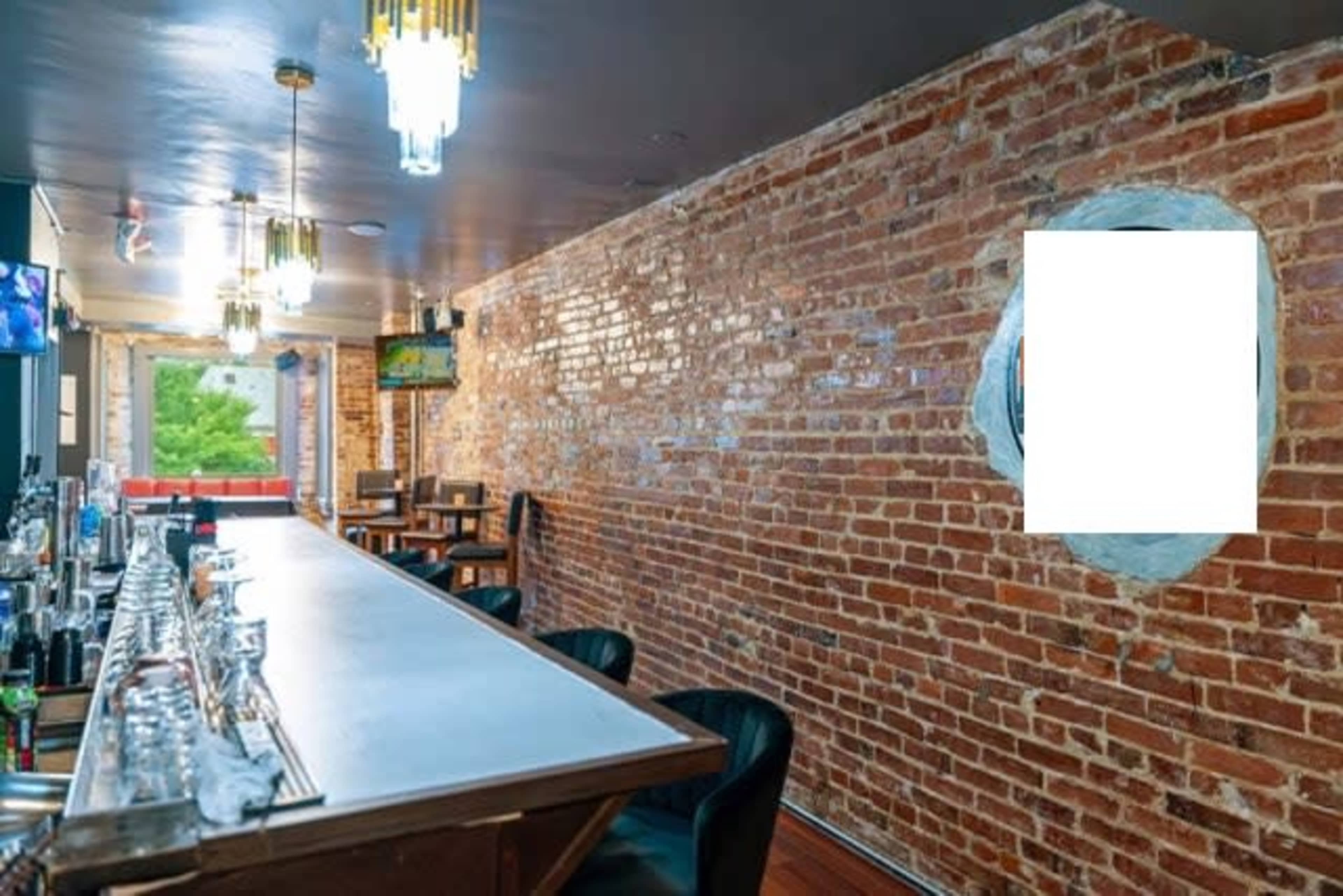 The image shows a bar area with a long countertop and exposed brick walls, leading to a window with a view of greenery outside.