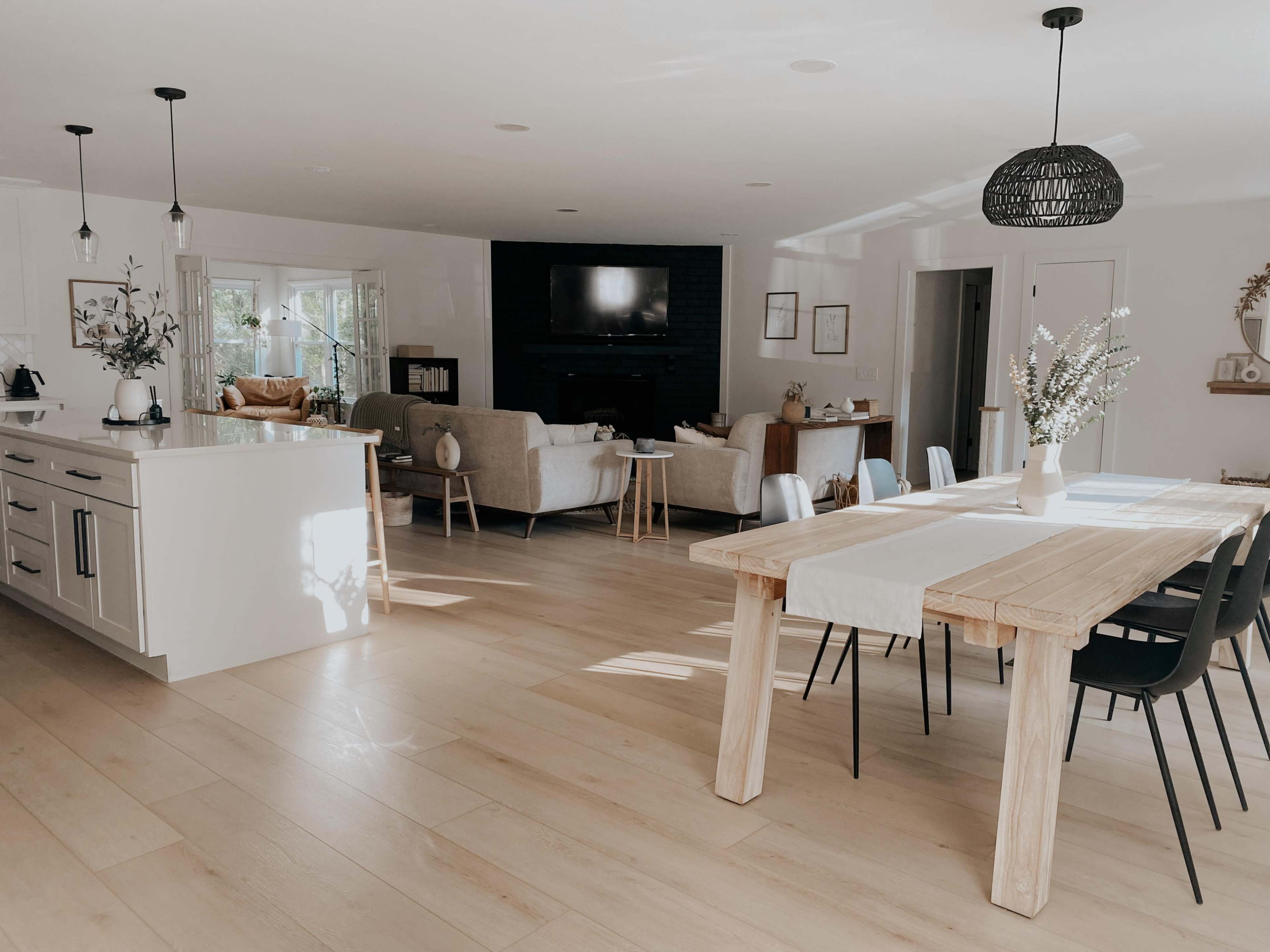 A modern open-concept living and dining area with a wooden table, black chairs, and a kitchen island, featuring light-colored flooring and natural light streaming through the windows.