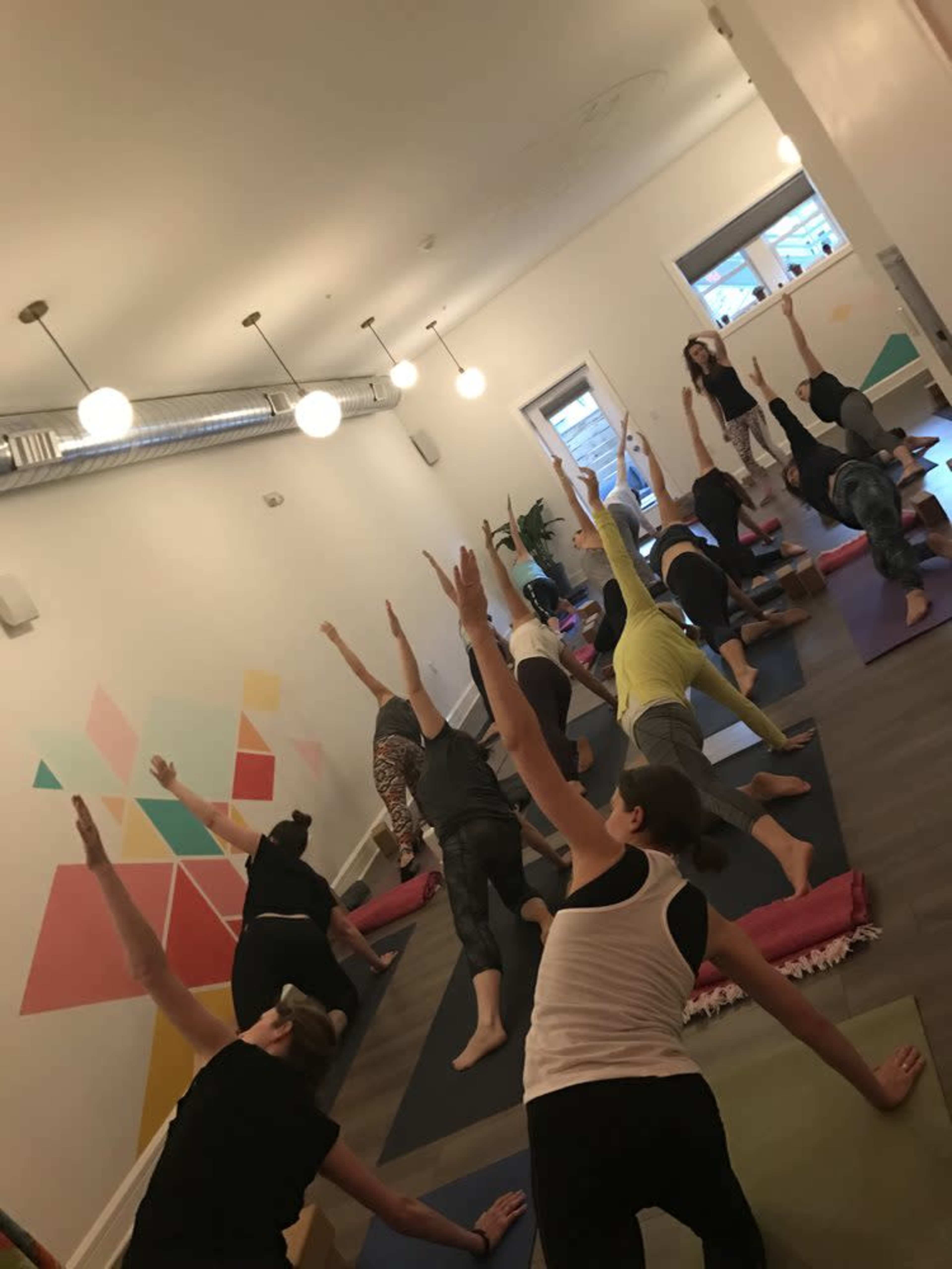 A group of people are practicing yoga in a brightly lit studio, with their arms extended towards the ceiling.