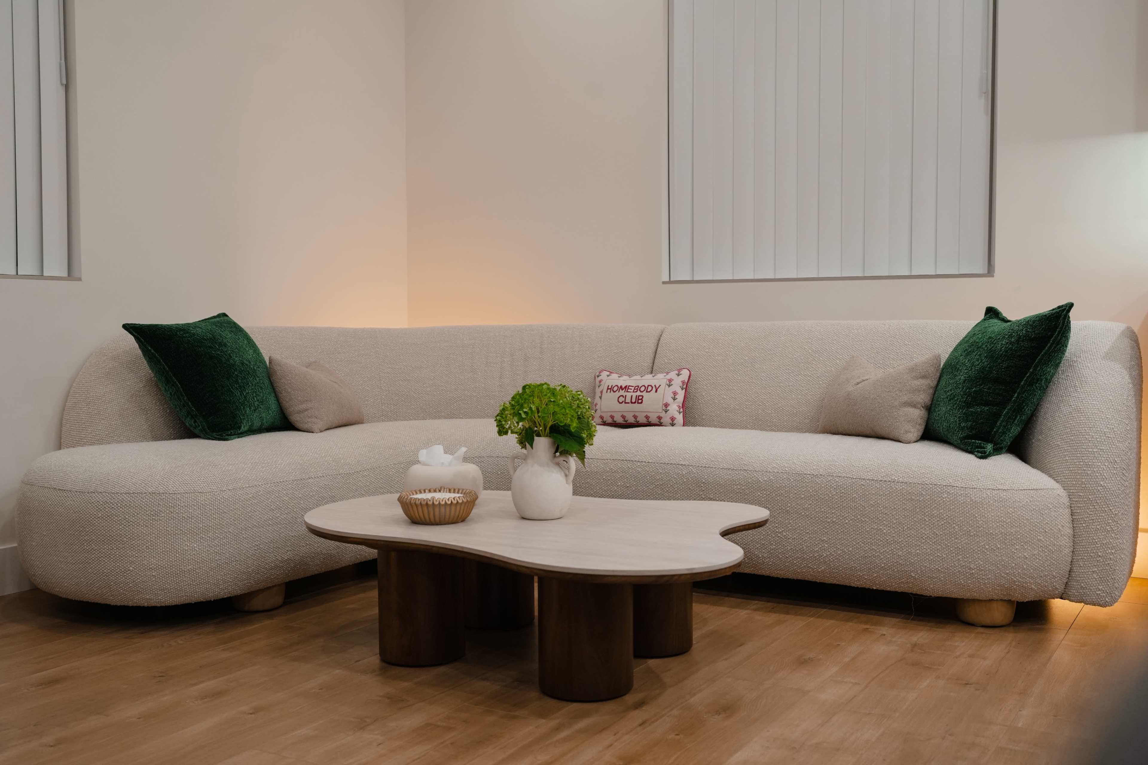The image shows a modern, curved sectional sofa with green cushions, accompanied by a round coffee table and a small potted plant in a light-colored room.