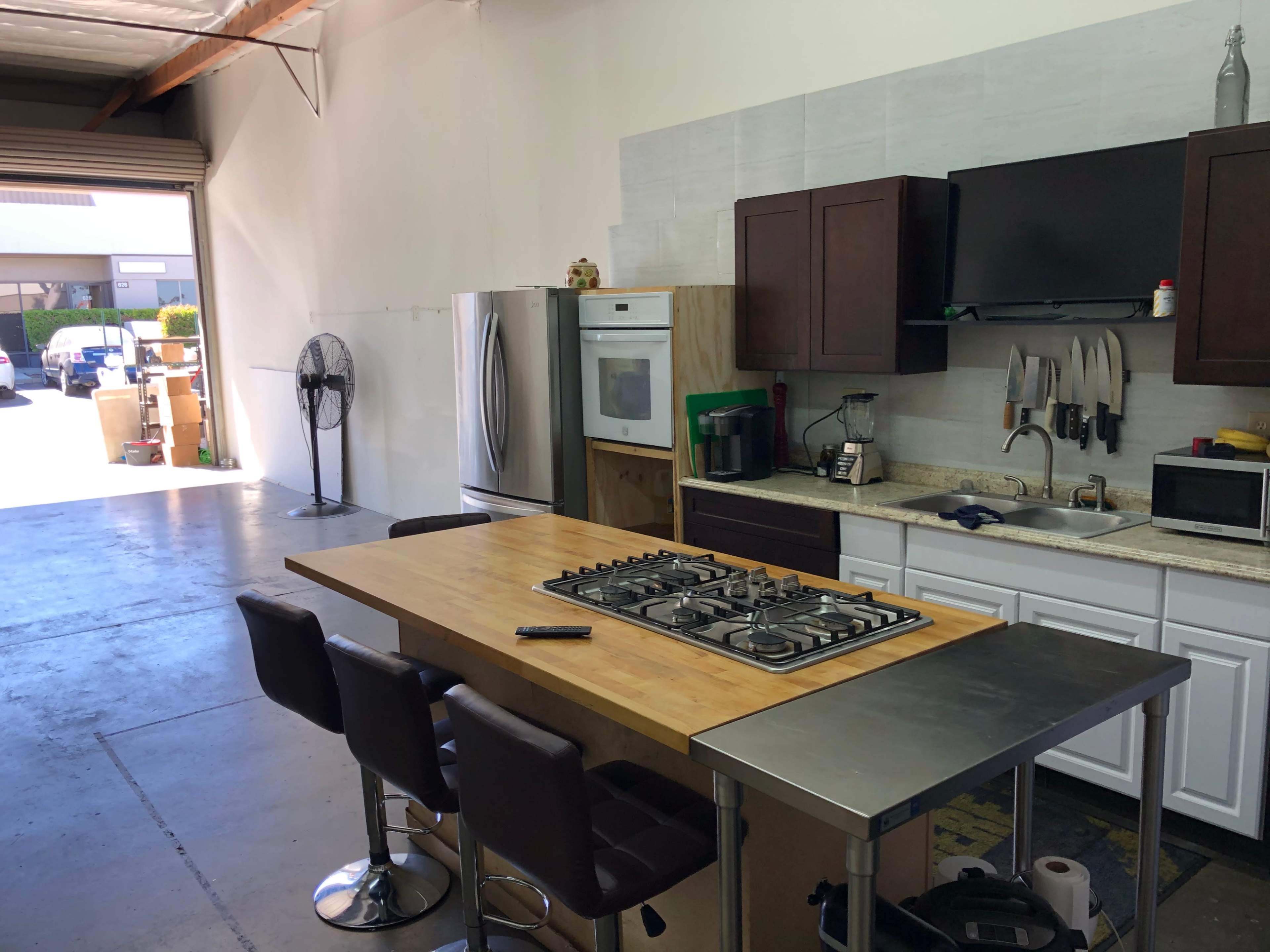 The image shows a spacious kitchen area in a warehouse, featuring a central island with a gas stove, stools, and appliances including a refrigerator and microwave.