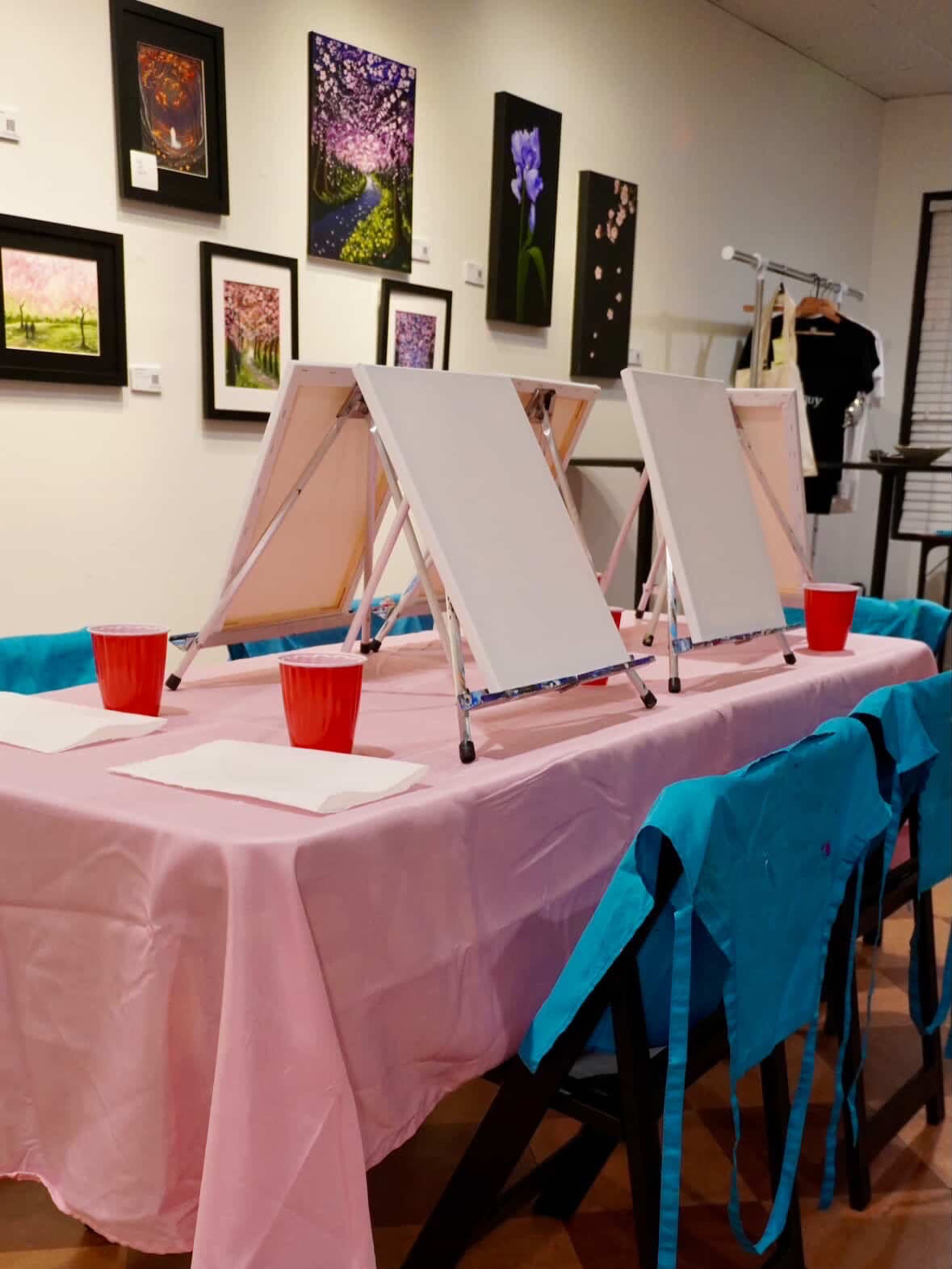 The image shows a table covered with a pink tablecloth, featuring three blank canvases on easels, red cups, and surrounded by blue chairs in an art studio setting.