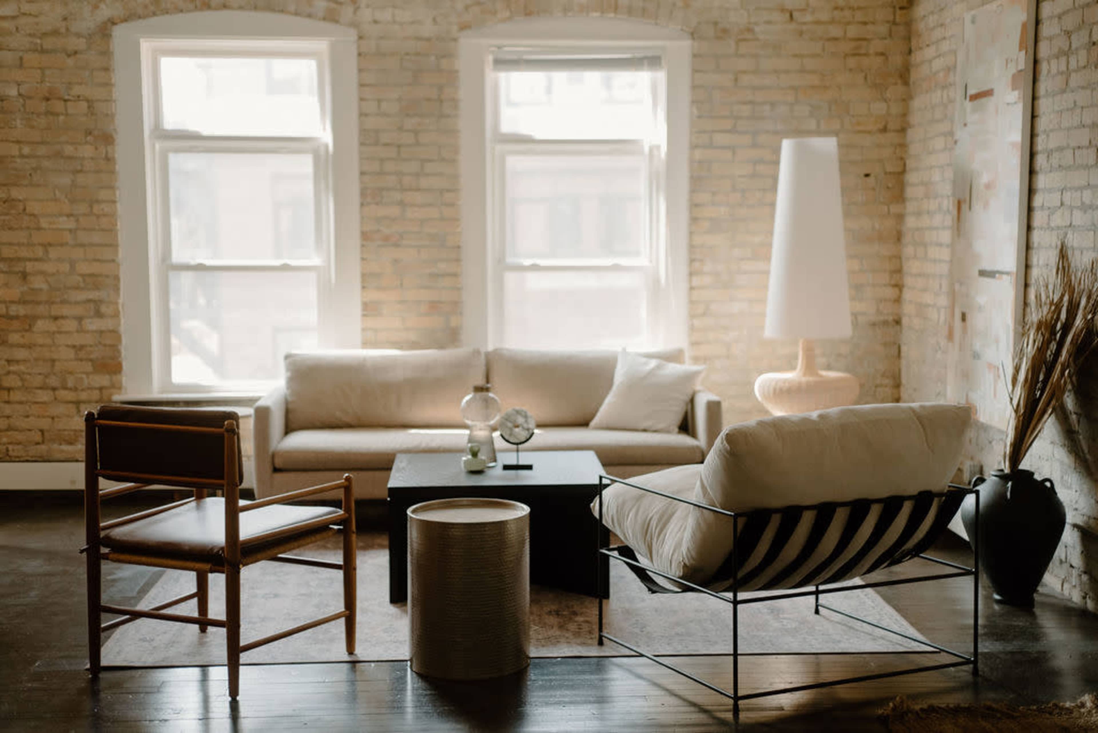 A stylish living room with a beige sofa, a black coffee table, and various decorative elements against a backdrop of exposed brick walls and large windows.