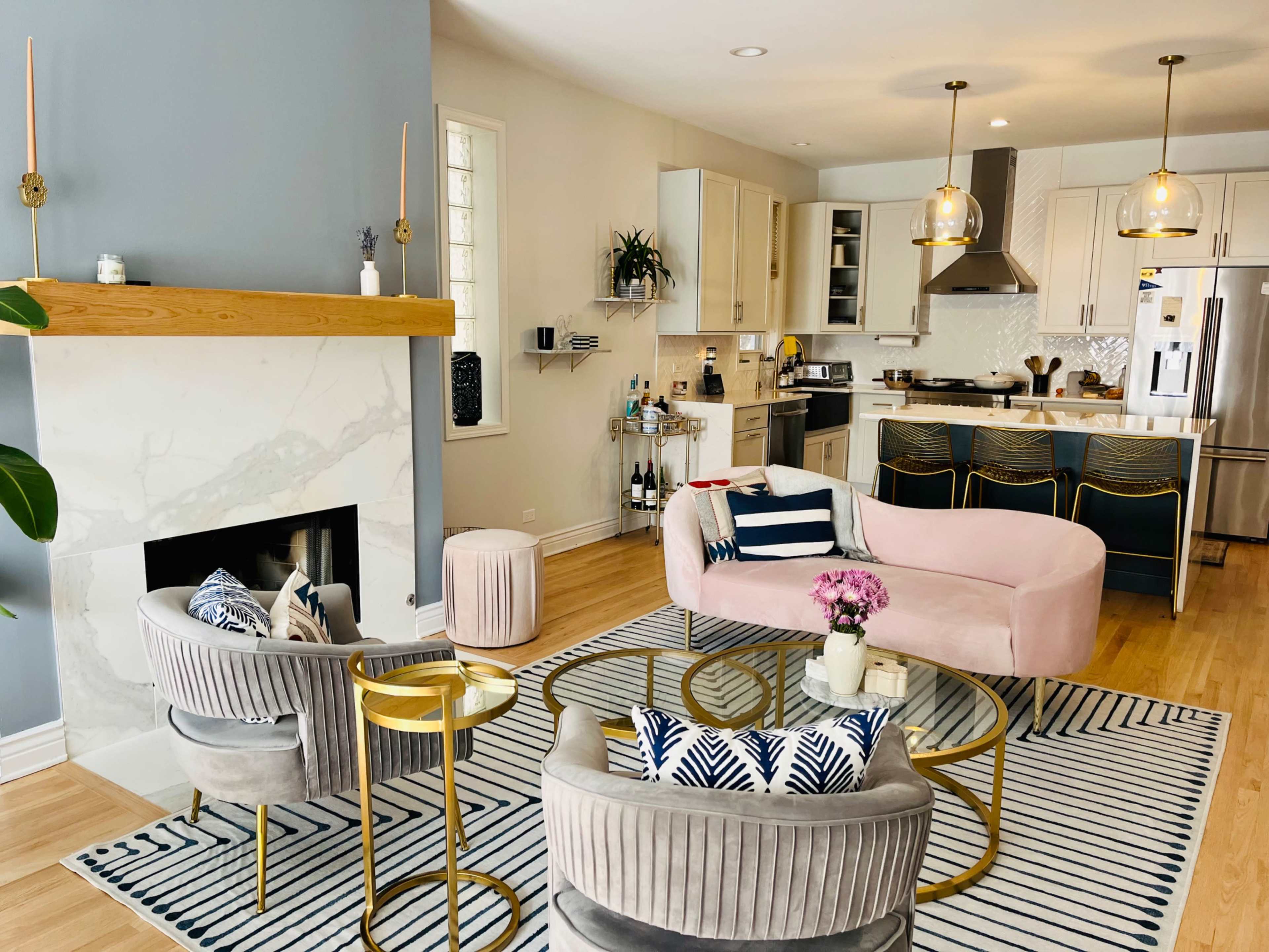 A modern living room with a pink sofa, two gray armchairs, a marble fireplace, and a kitchen area in the background.