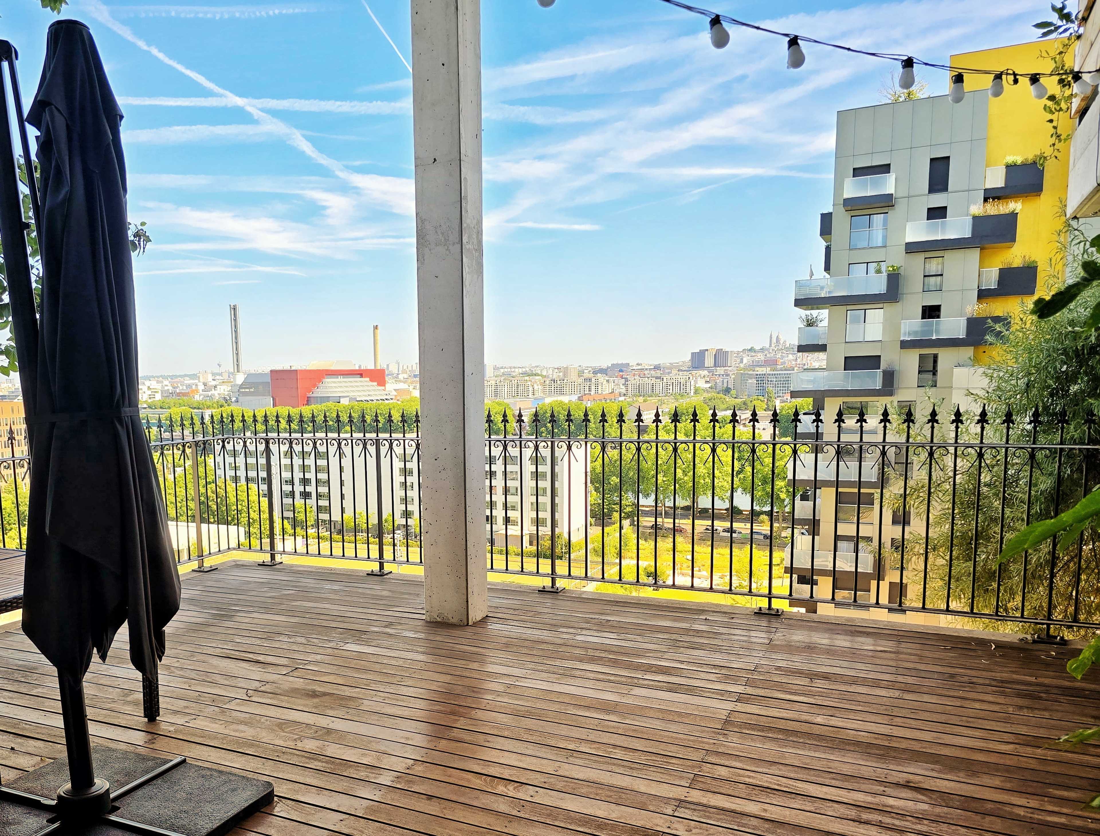 A wooden deck with a standing umbrella overlooks a cityscape featuring buildings and distant industrial structures under a clear blue sky.