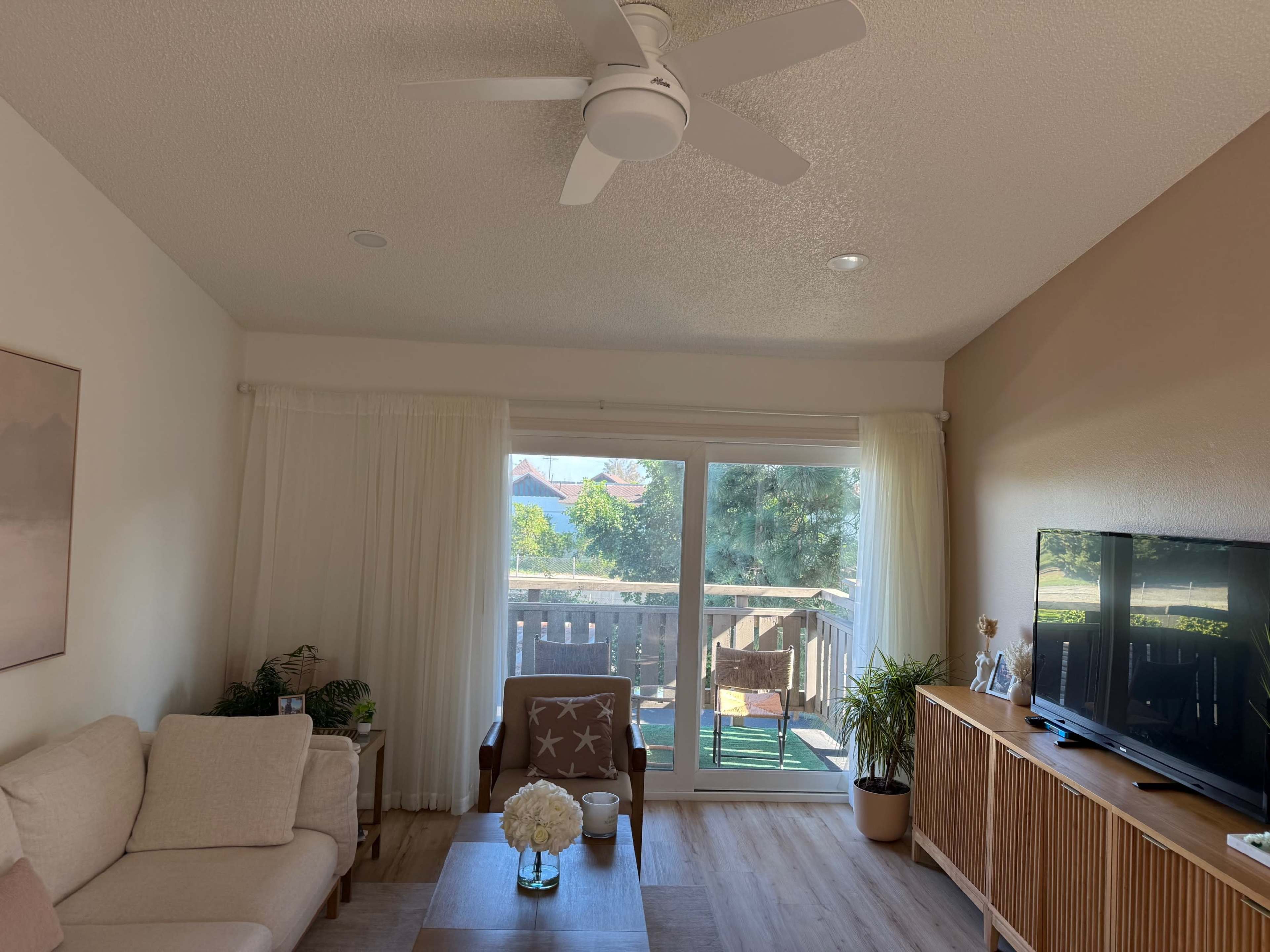 The image shows a living room with a white couch, a wooden coffee table, a television on a cabinet, and large windows that overlook a balcony with greenery.