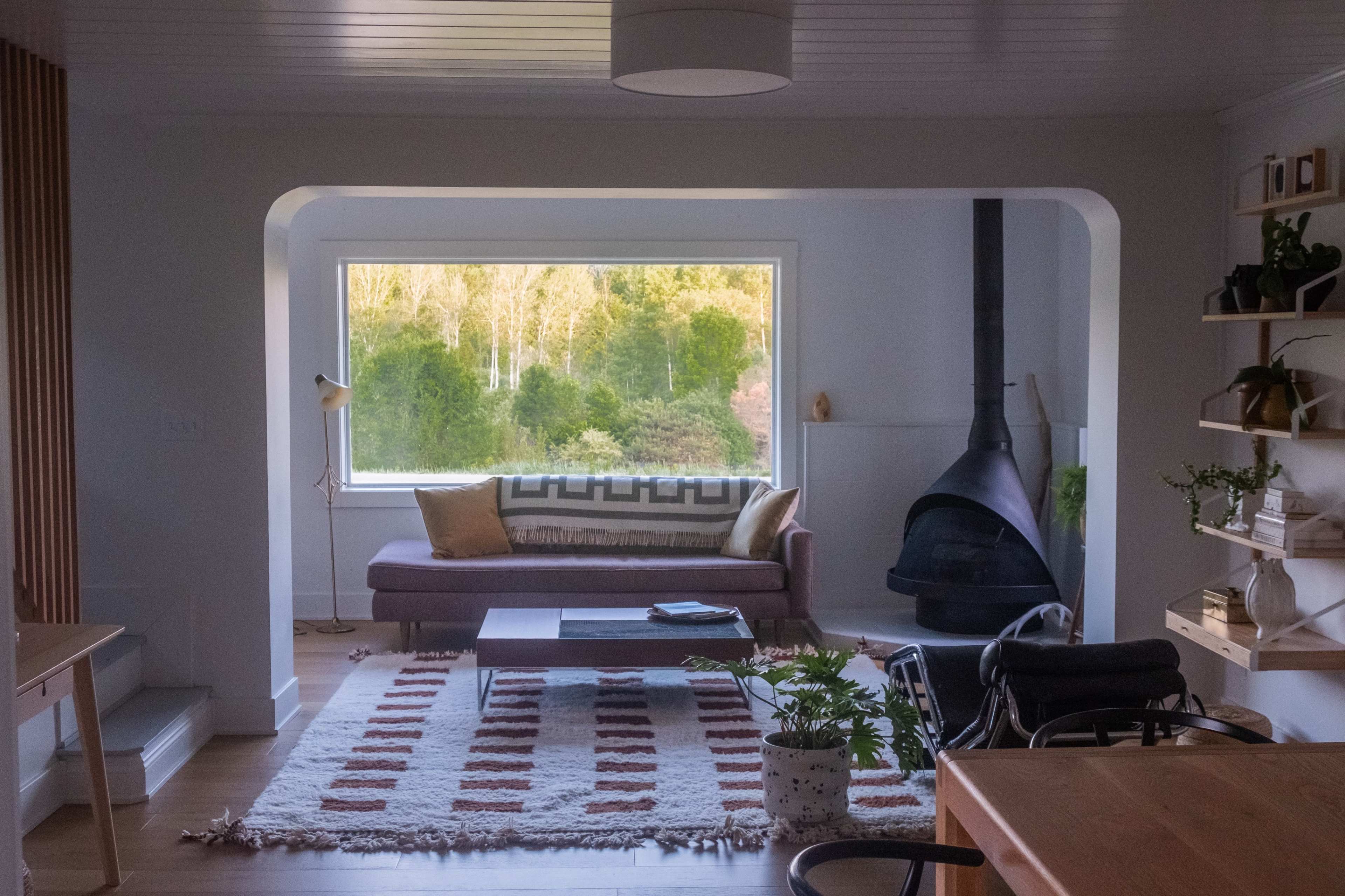 A living room with a large window overlooking a wooded area, a brown sofa, a coffee table, and a black fireplace.