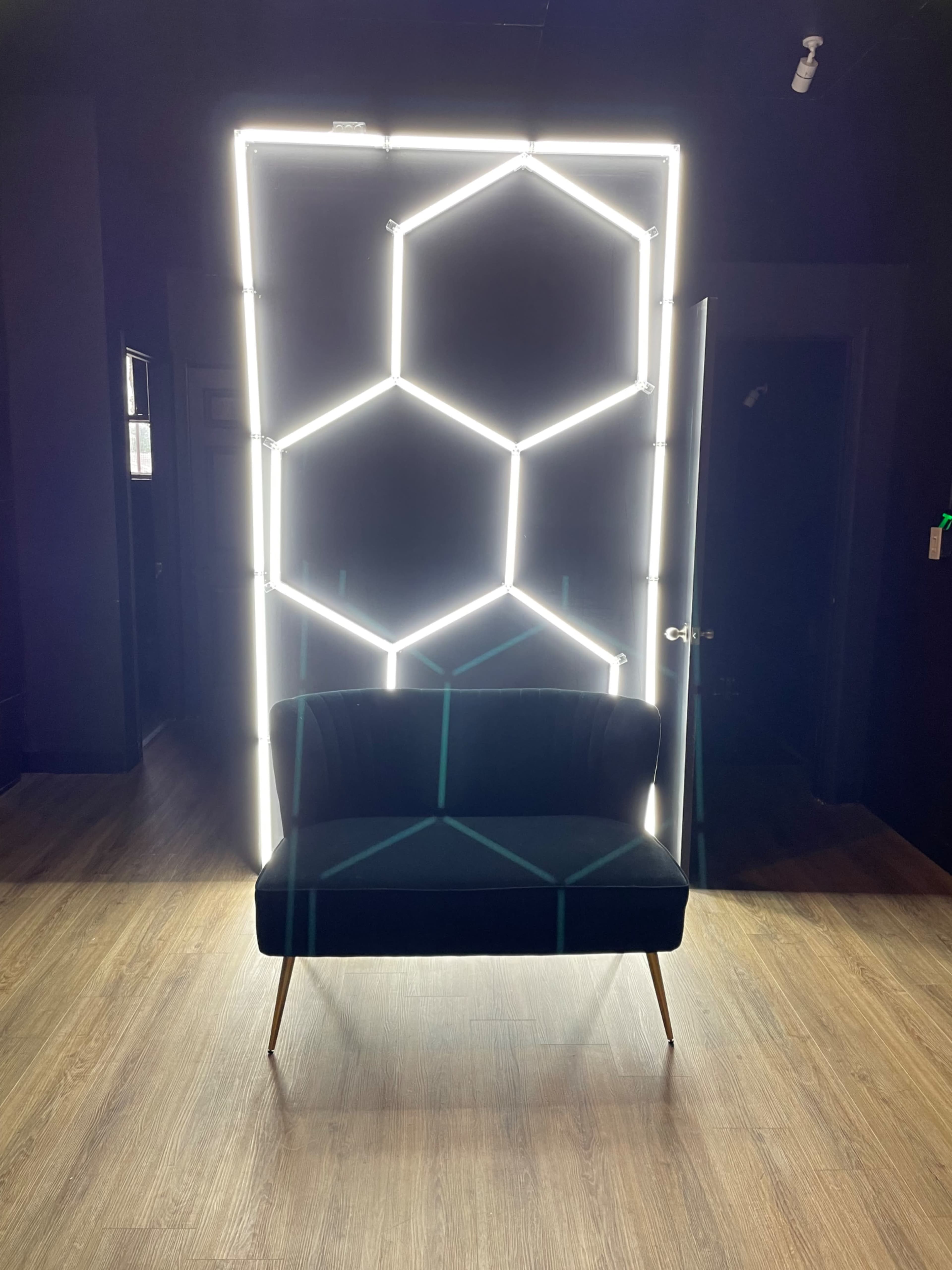 A black velvet couch is positioned in front of a neon-lit hexagonal patterned backdrop in a dimly lit room.