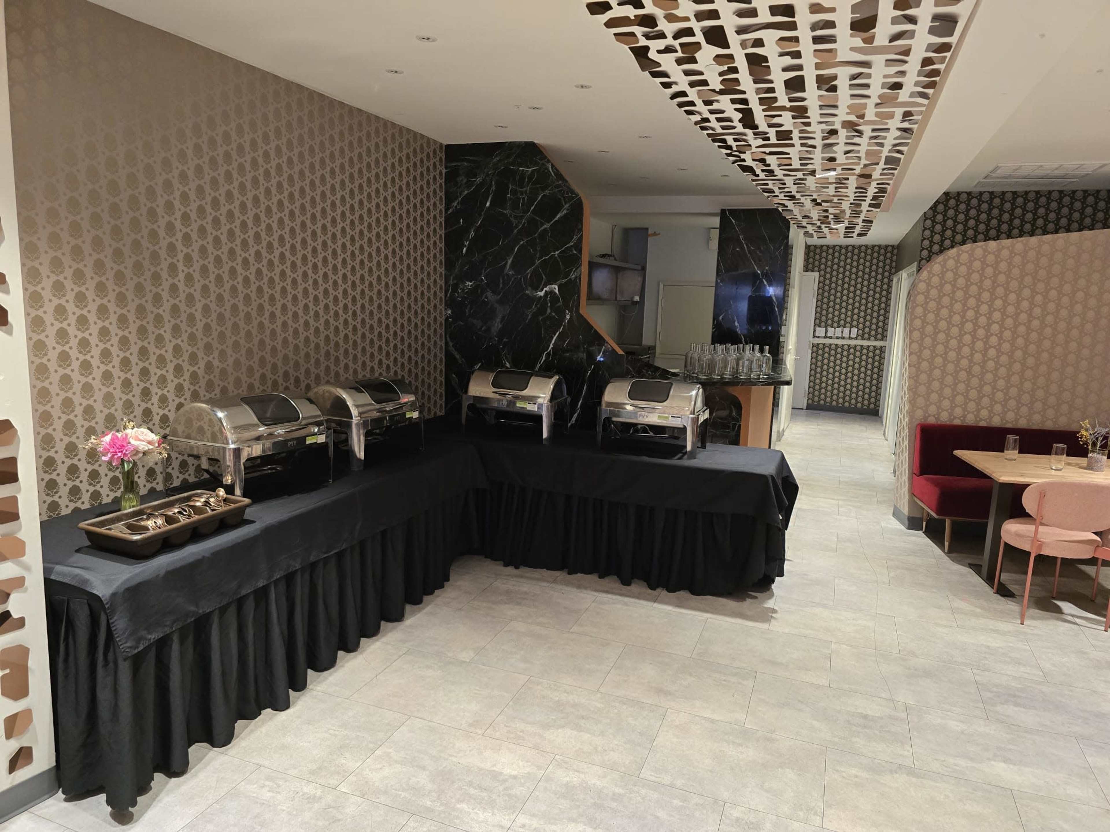 The image shows a dining area with several chafing dishes on a black tablecloth, a marble wall, and a seating area with chairs and tables.