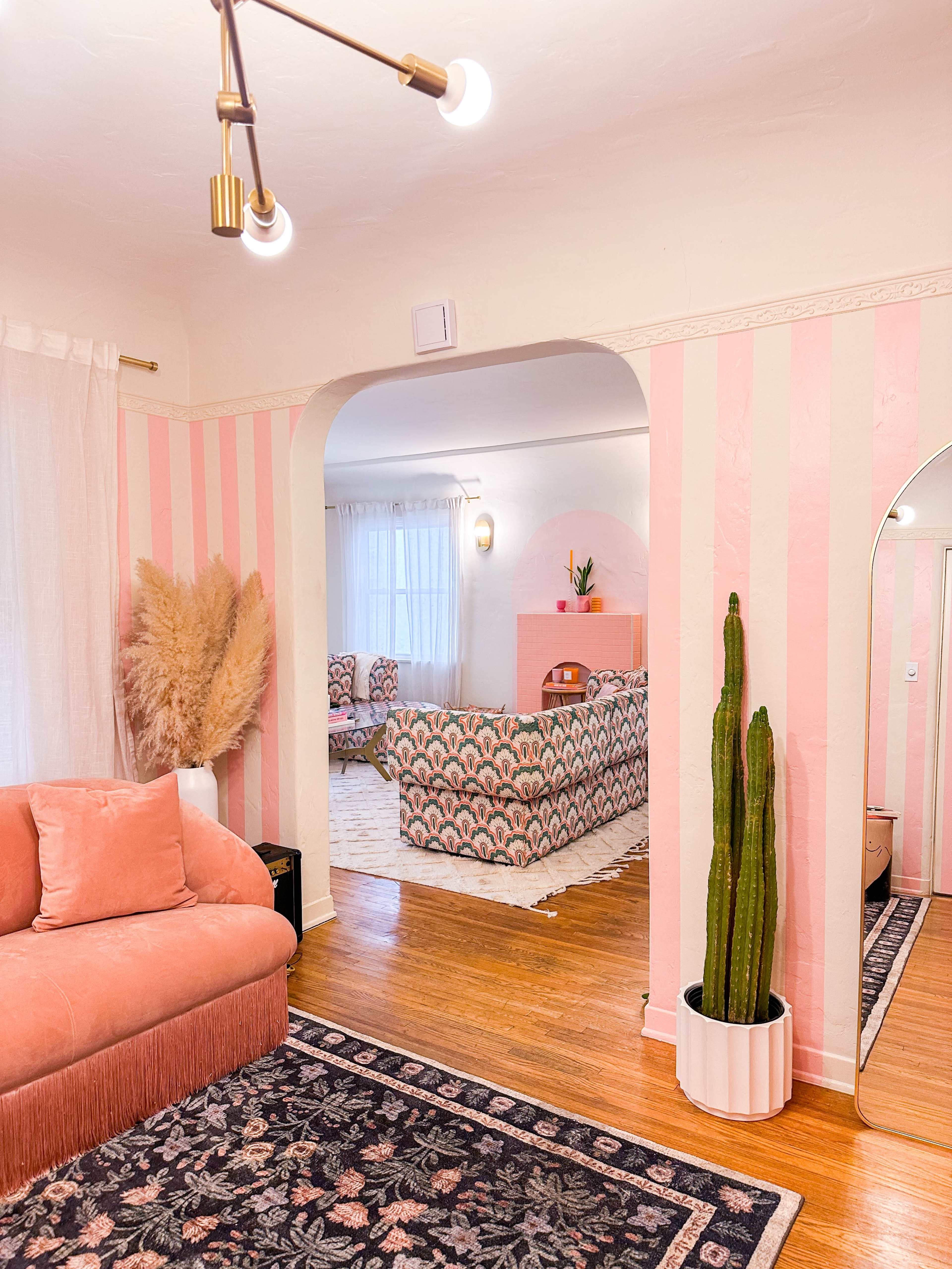 The image shows a living room featuring striped pink and white walls, a pink archway, a cactus in a white pot, and two seating areas, one with a plush sofa and another with patterned chairs.