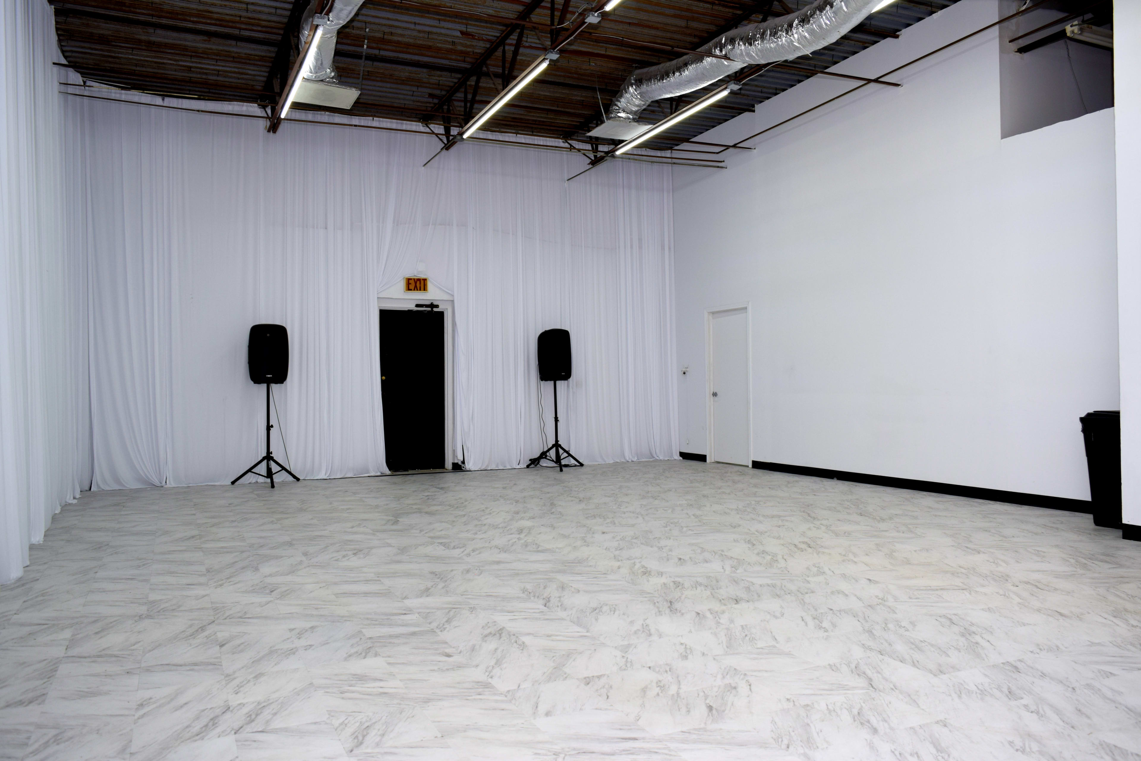 The image shows a spacious, empty room with white walls, a marble-patterned floor, two sound speakers on stands, and a closed door at the back.