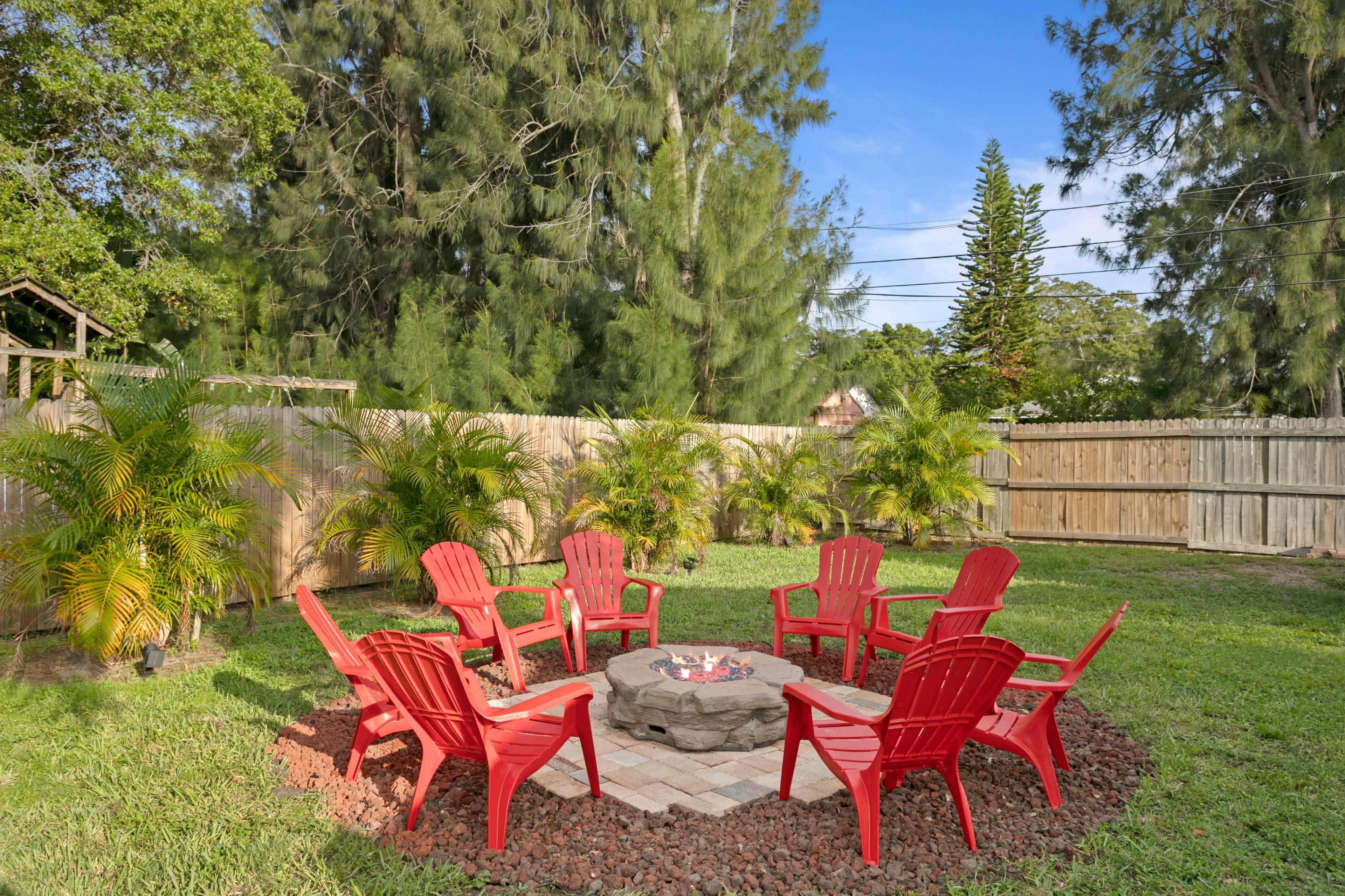 A circular seating arrangement of red chairs surrounds a stone fire pit in a yard with green grass and trees.