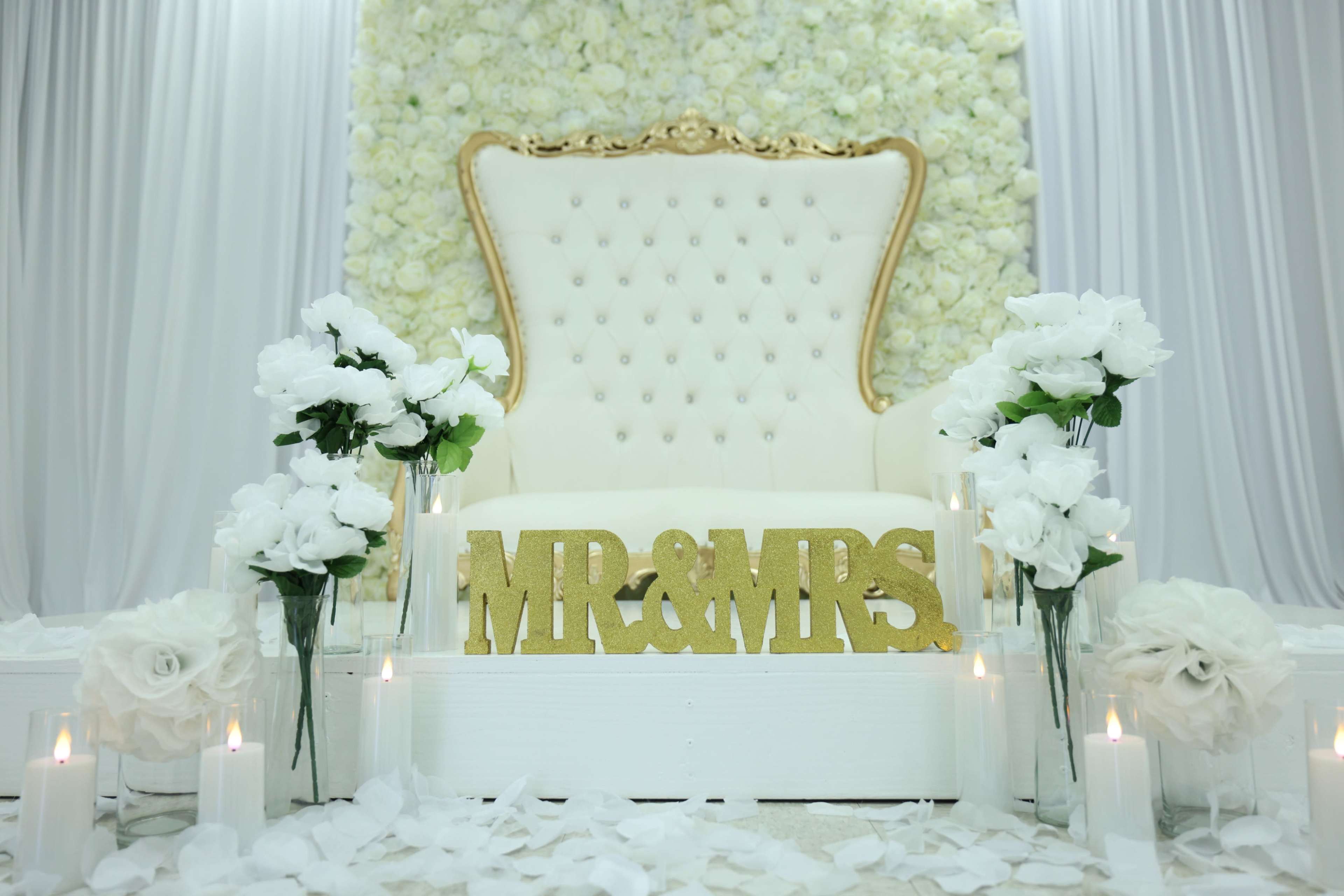 A white tufted throne with a gold frame is set against a backdrop of artificial flowers, accompanied by floral arrangements and candles on each side.