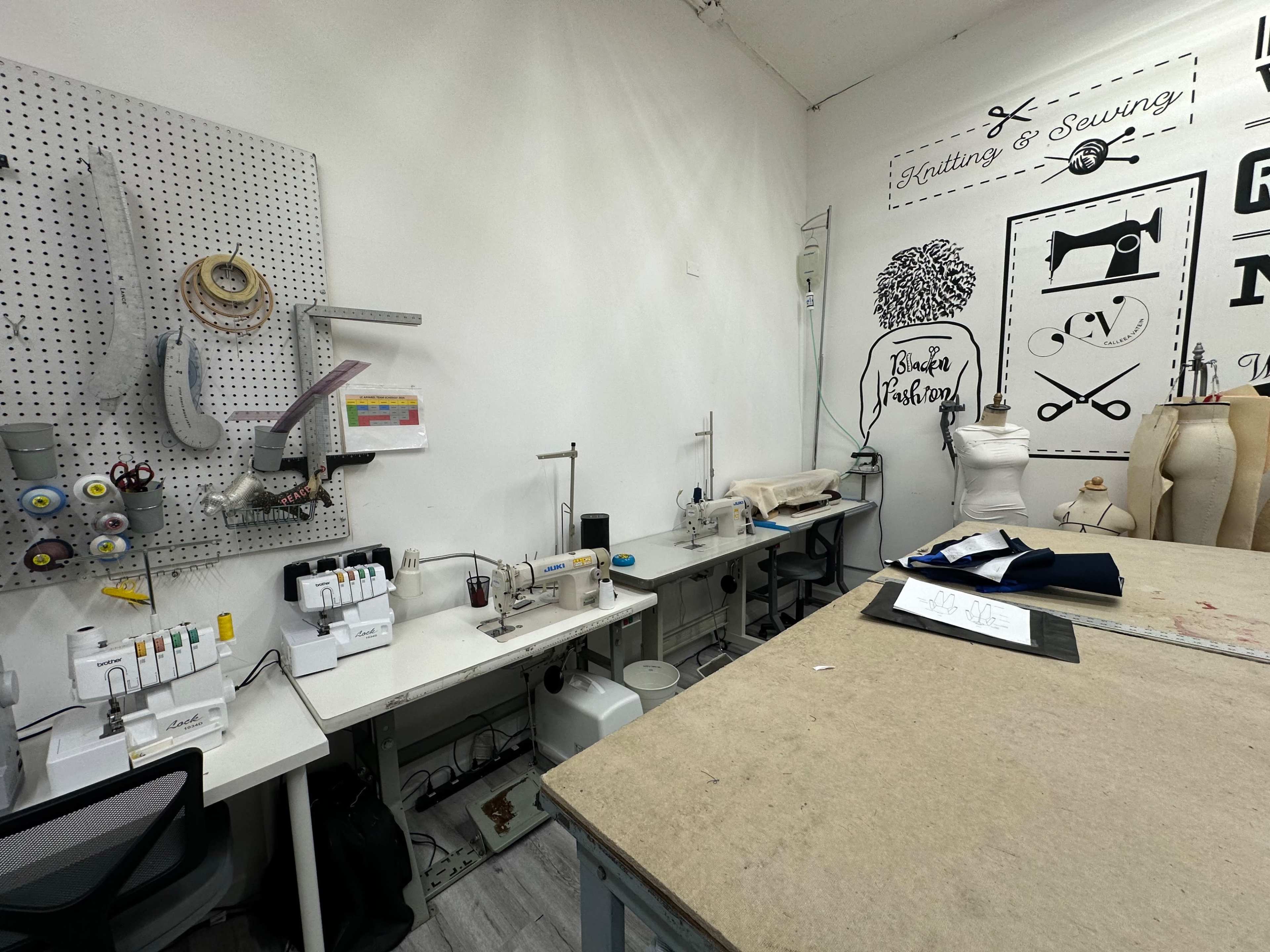 A sewing workshop with several sewing machines, a cutting table, and a wall decorated with sewing-related graphics.