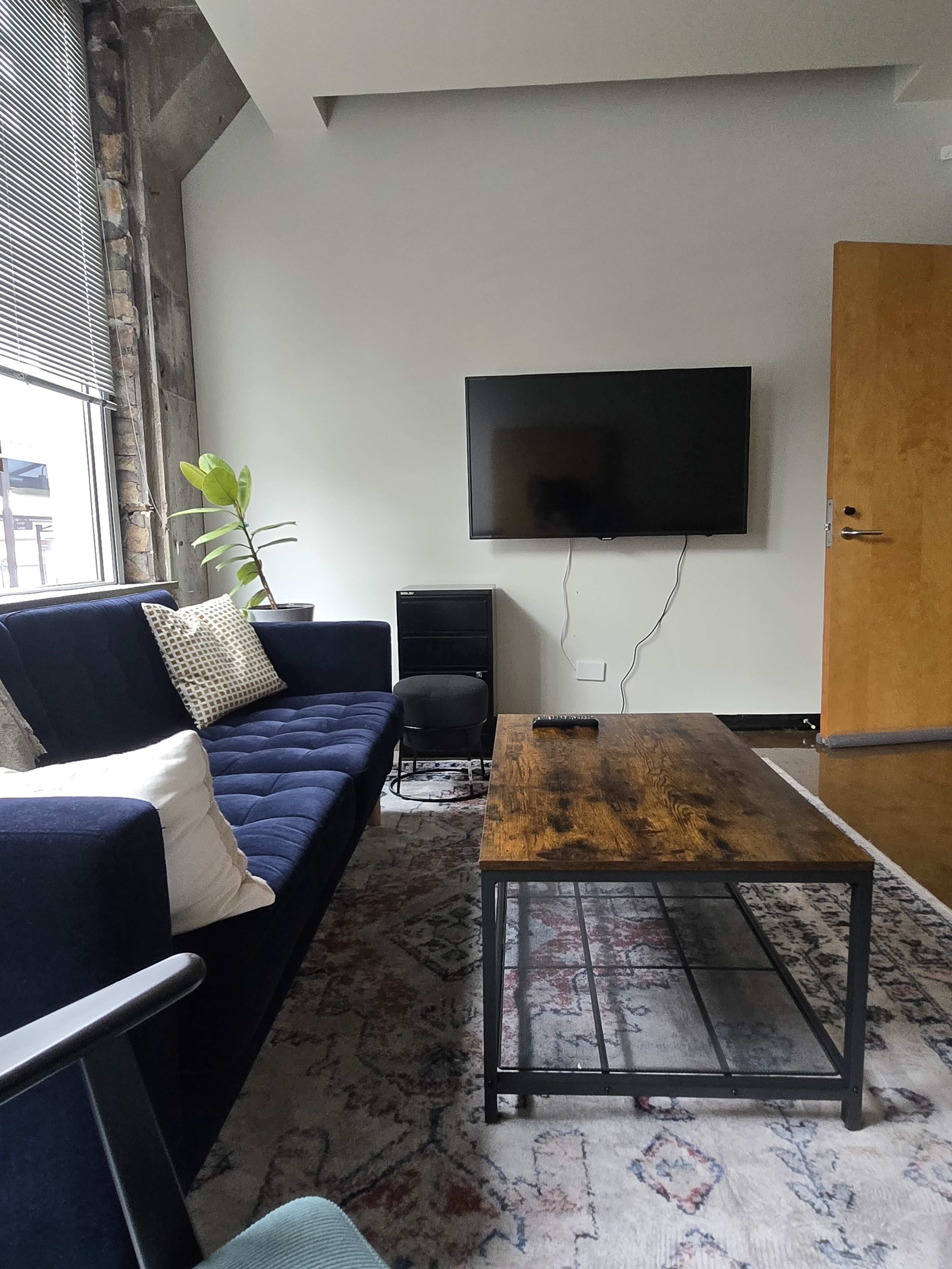 The image shows a modern living room with a blue couch, a wooden coffee table, a flat-screen TV mounted on the wall, and a green plant near the window.