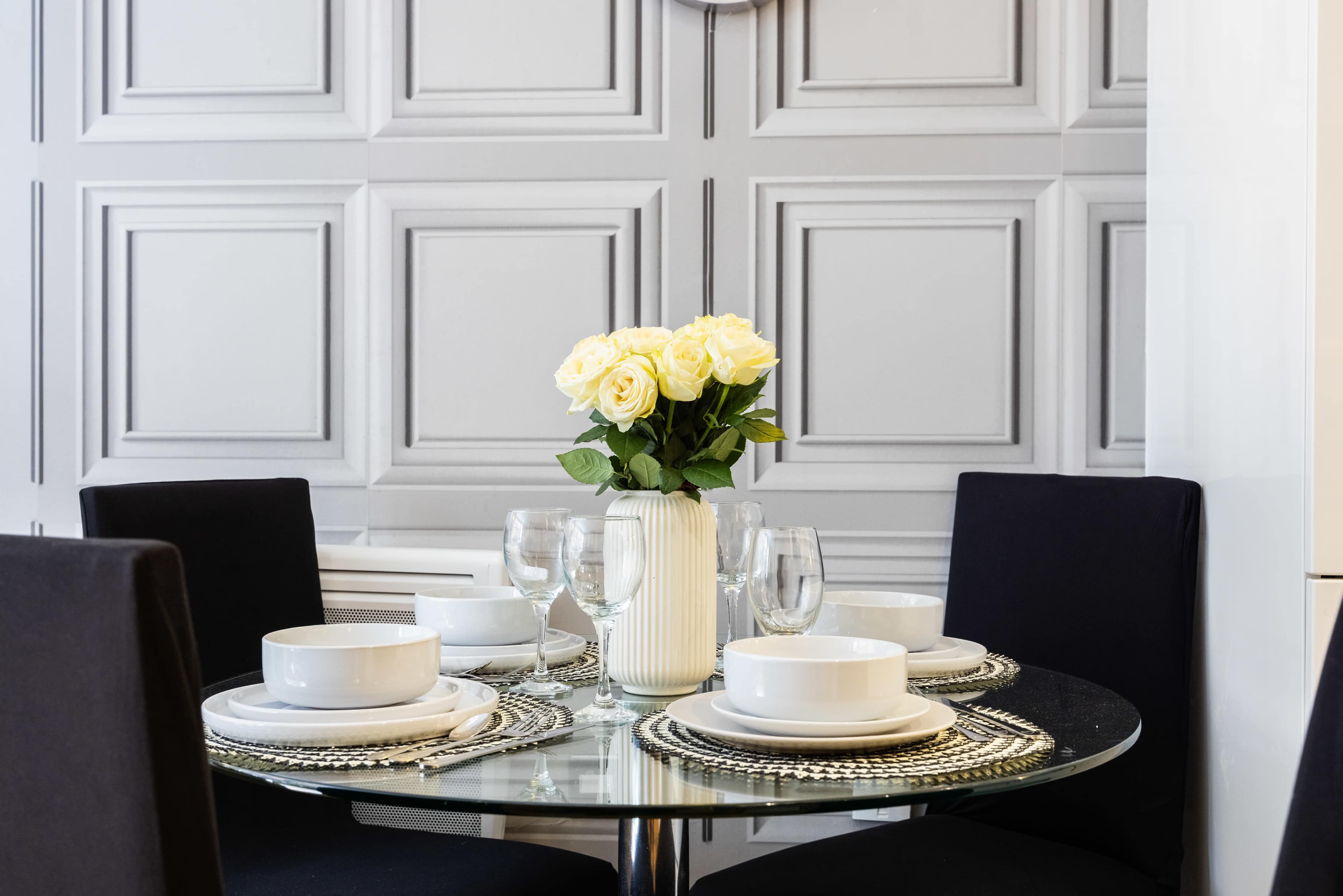 A round glass dining table is set with white plates and transparent glasses, accompanied by a vase of yellow roses in a room with textured gray walls.