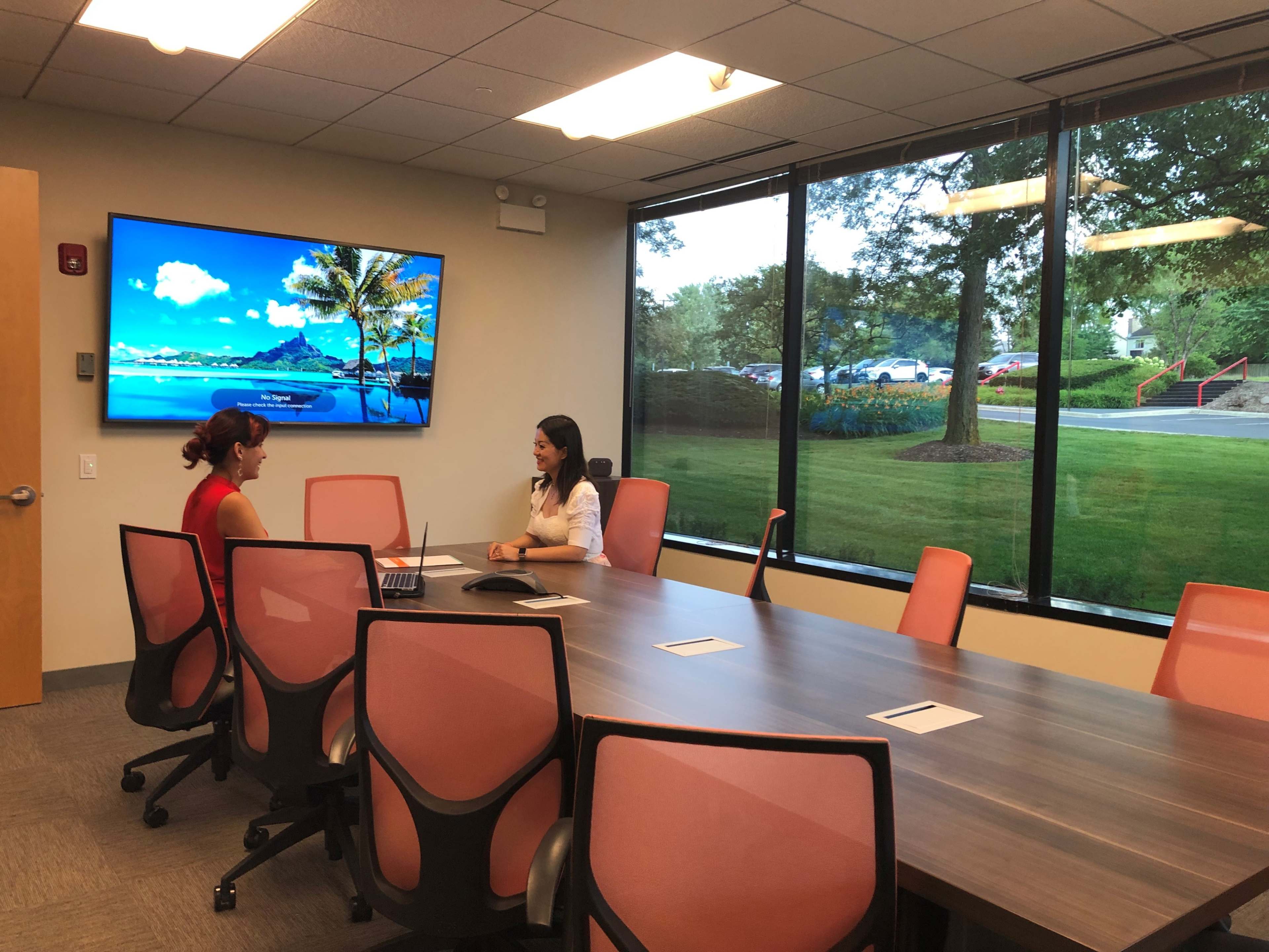 Two women are seated at a conference table in a well-lit meeting room, with a large-screen display showing a beach scene in the background and windows overlooking a green landscape.