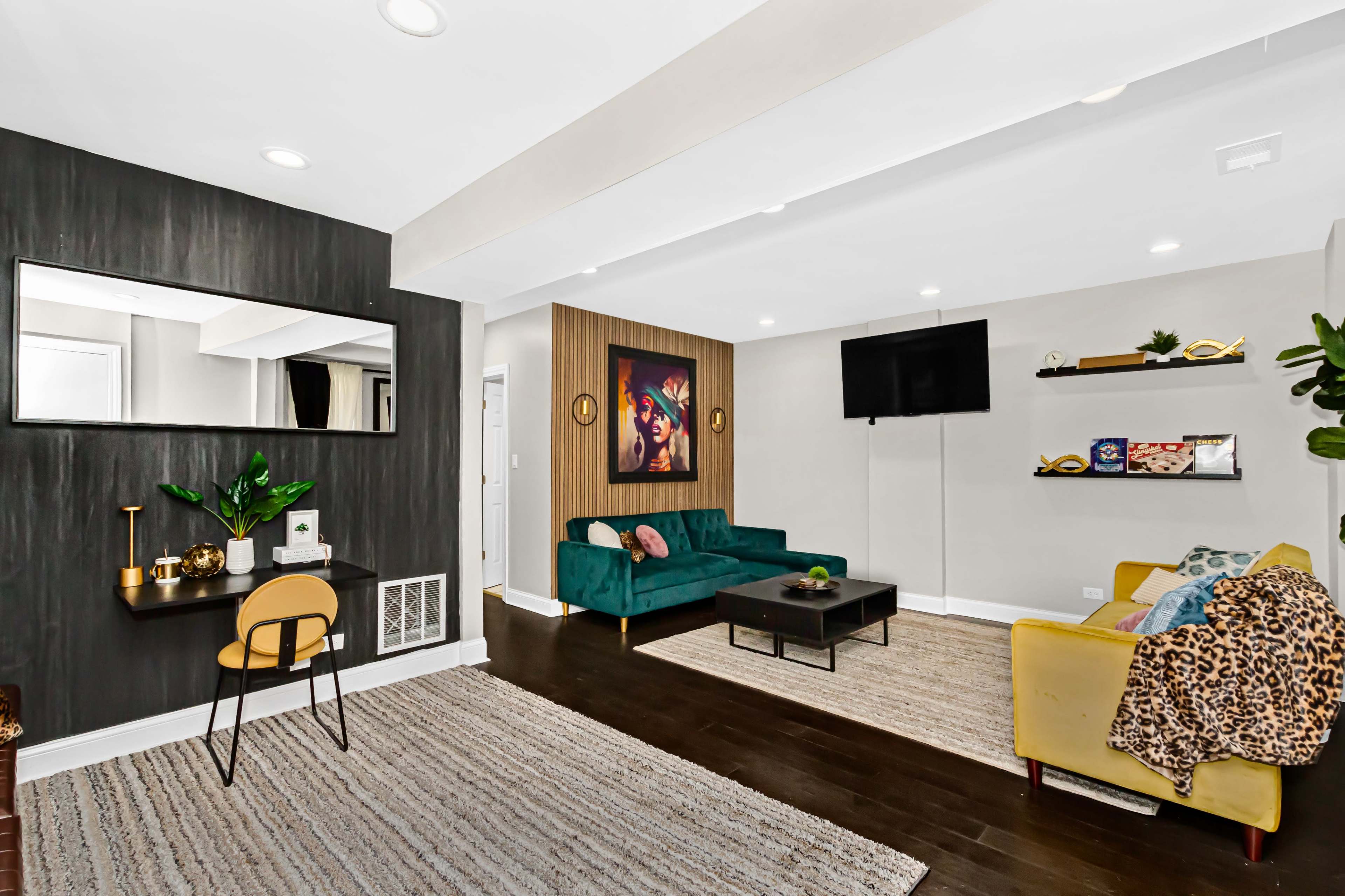 A modern living room featuring a green sofa, a yellow armchair, a black coffee table, a television mounted on the wall, and artwork on display.