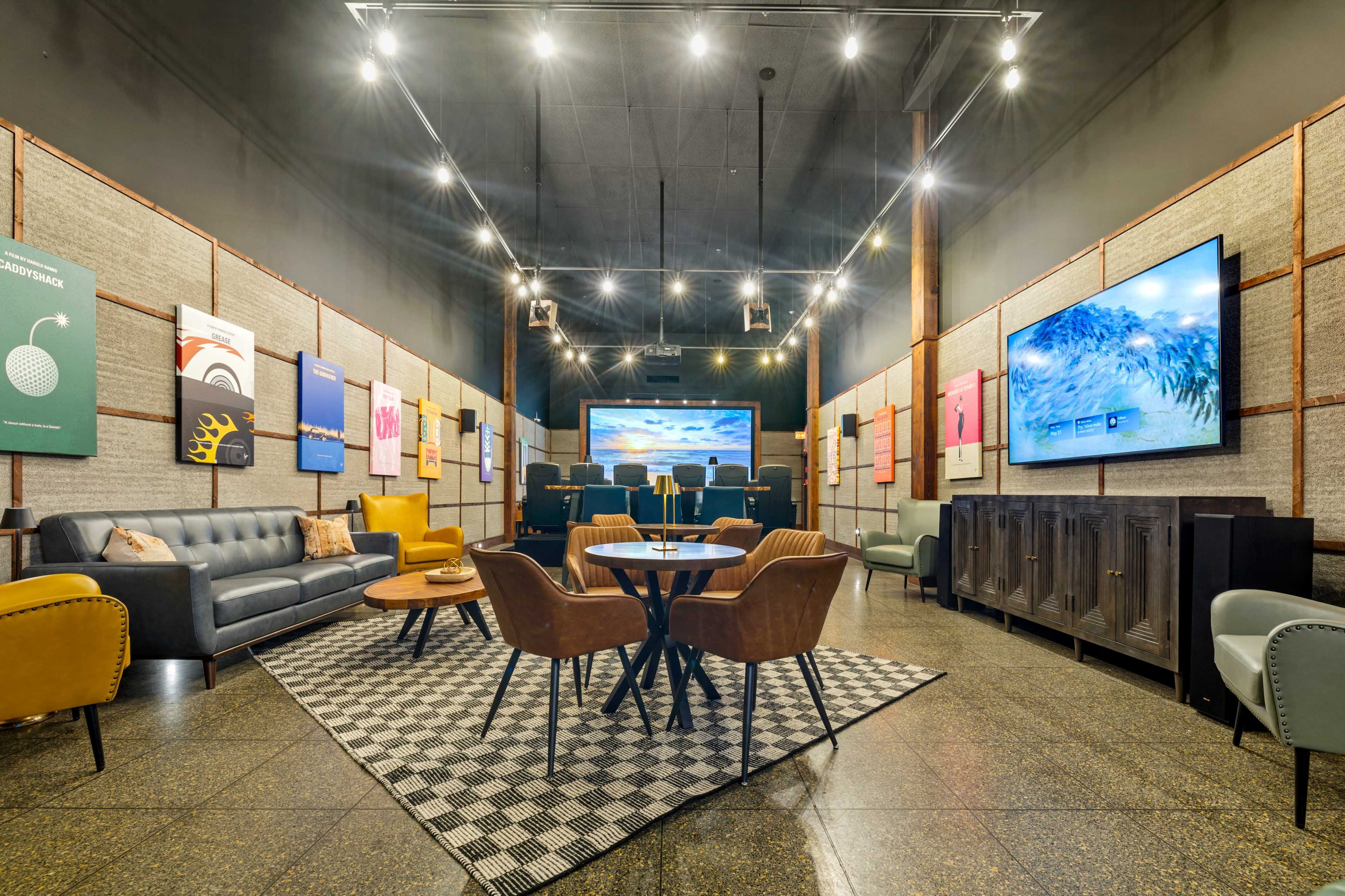 The image depicts a stylish lounge with a cozy seating area featuring a gray sofa, multiple armchairs, a round table, and large screens displaying artwork.
