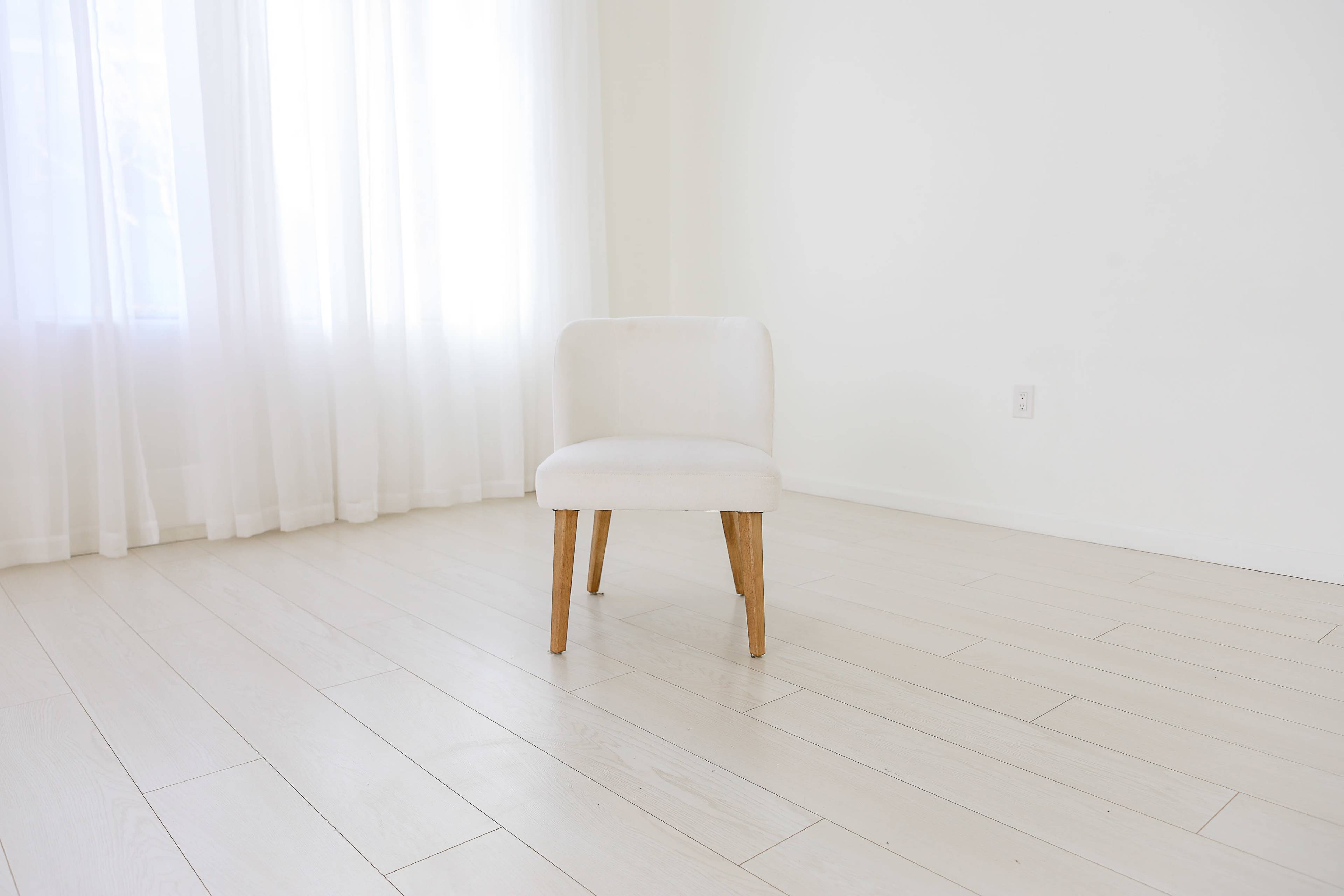 A single white chair stands on a light-colored wooden floor in a bright, minimalist room with sheer white curtains.