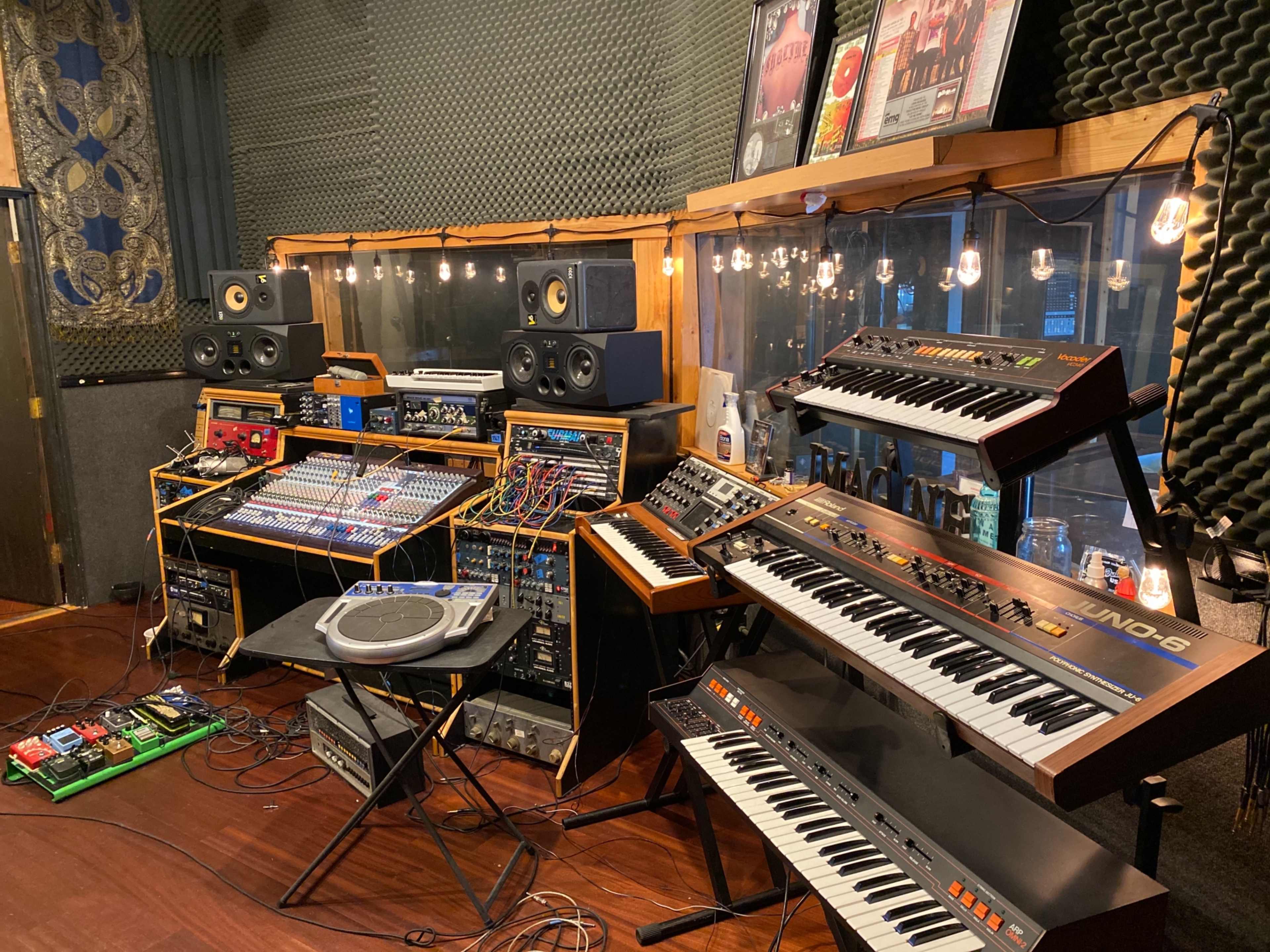 A music studio with various synthesizers, a mixing console, and studio monitors arranged around the space.