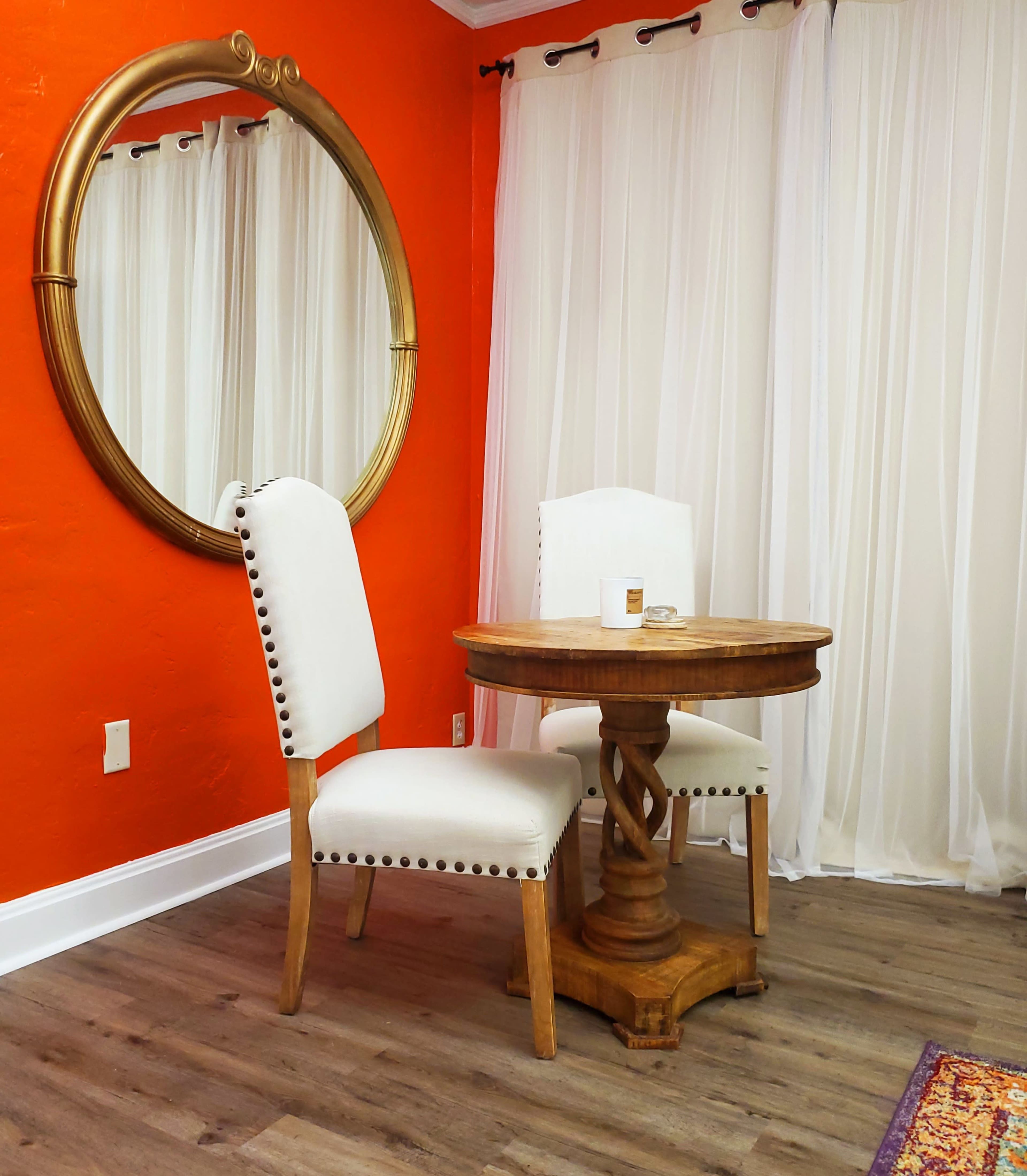 A round wooden table with two upholstered chairs is set against an orange wall, with a large round mirror hanging above.